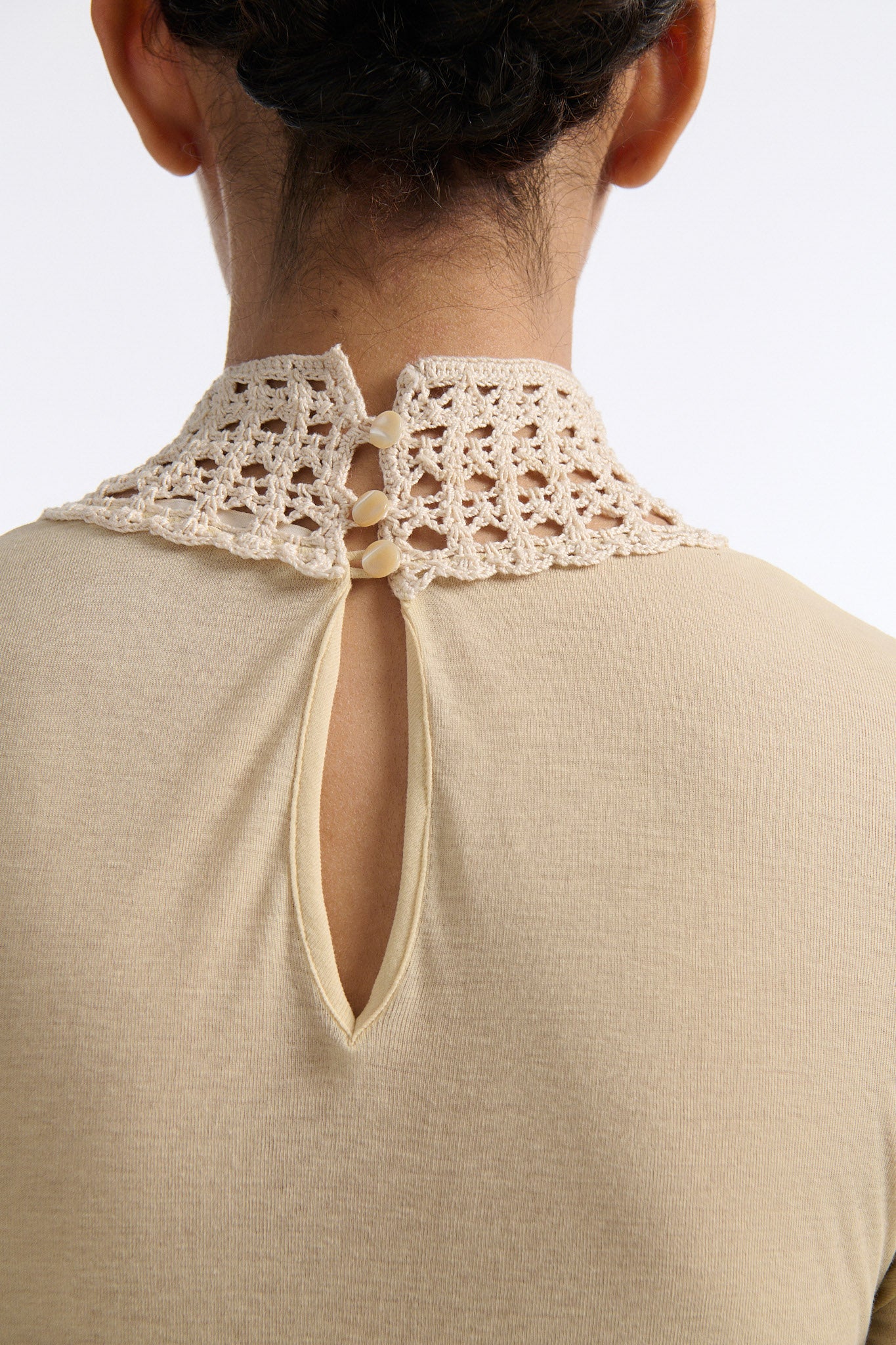 Female model with slicked back black hair in a bun wears beige linen handkerchief wrap skirt. Waist and closure are lined with melted metallic embellishments in gold and silver to provide an oyster illusion. Model also wears light beige fine-gauge cotton long sleeve top with mockneck crochet collar.