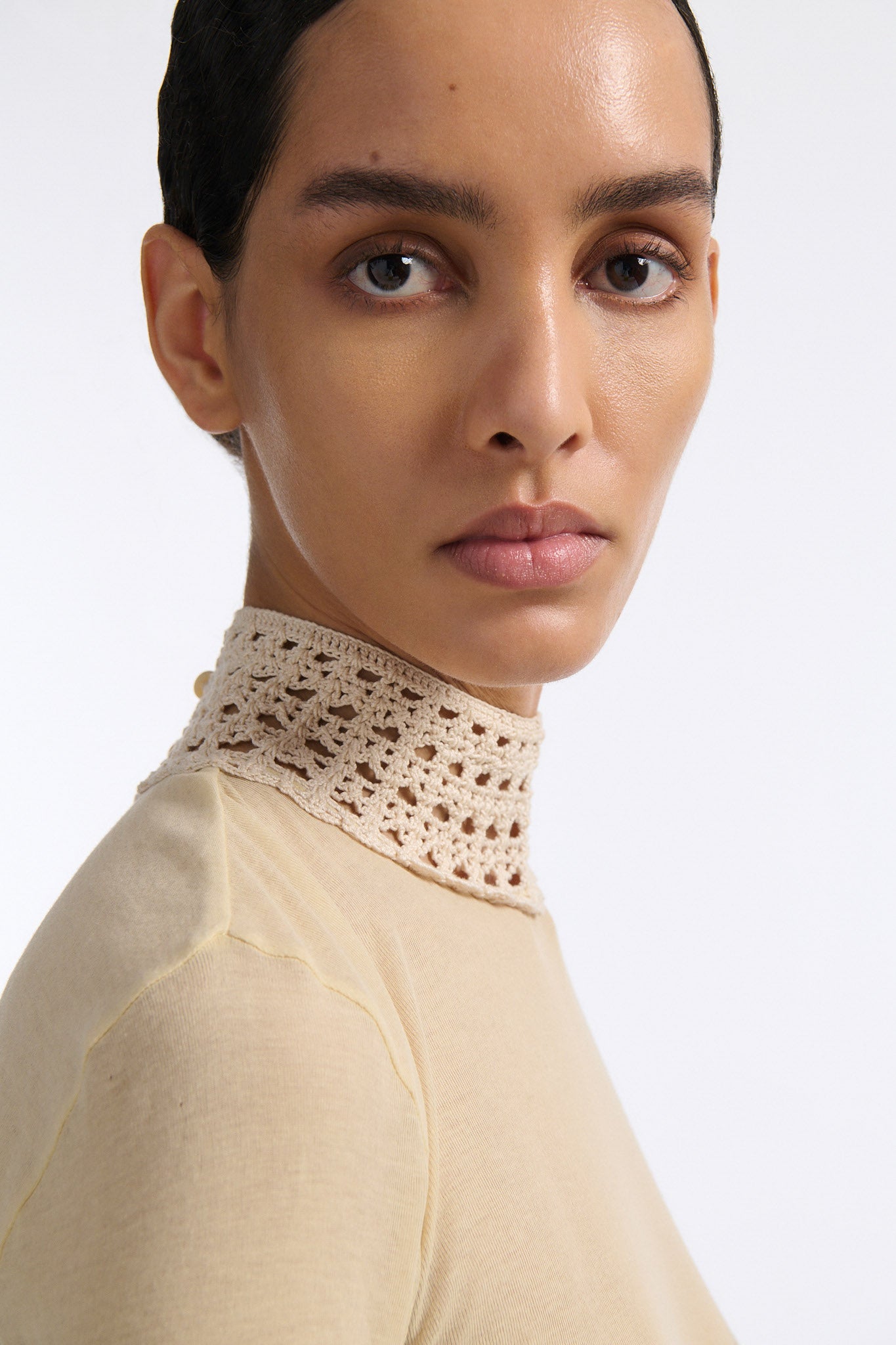 Female model with slicked back black hair in a bun wears beige linen handkerchief wrap skirt. Waist and closure are lined with melted metallic embellishments in gold and silver to provide an oyster illusion. Model also wears light beige fine-gauge cotton long sleeve top with mockneck crochet collar.