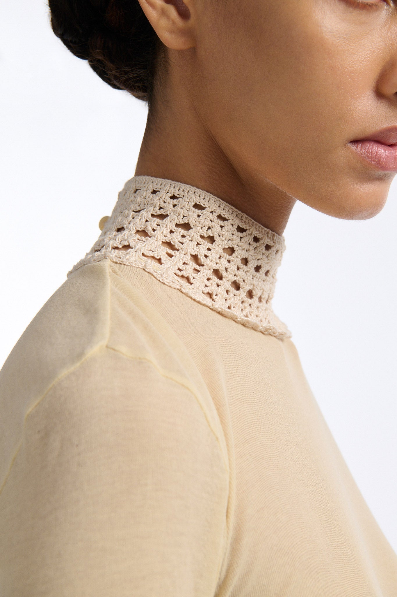 Female model with slicked back black hair in a bun wears beige linen handkerchief wrap skirt. Waist and closure are lined with melted metallic embellishments in gold and silver to provide an oyster illusion. Model also wears light beige fine-gauge cotton long sleeve top with mockneck crochet collar.