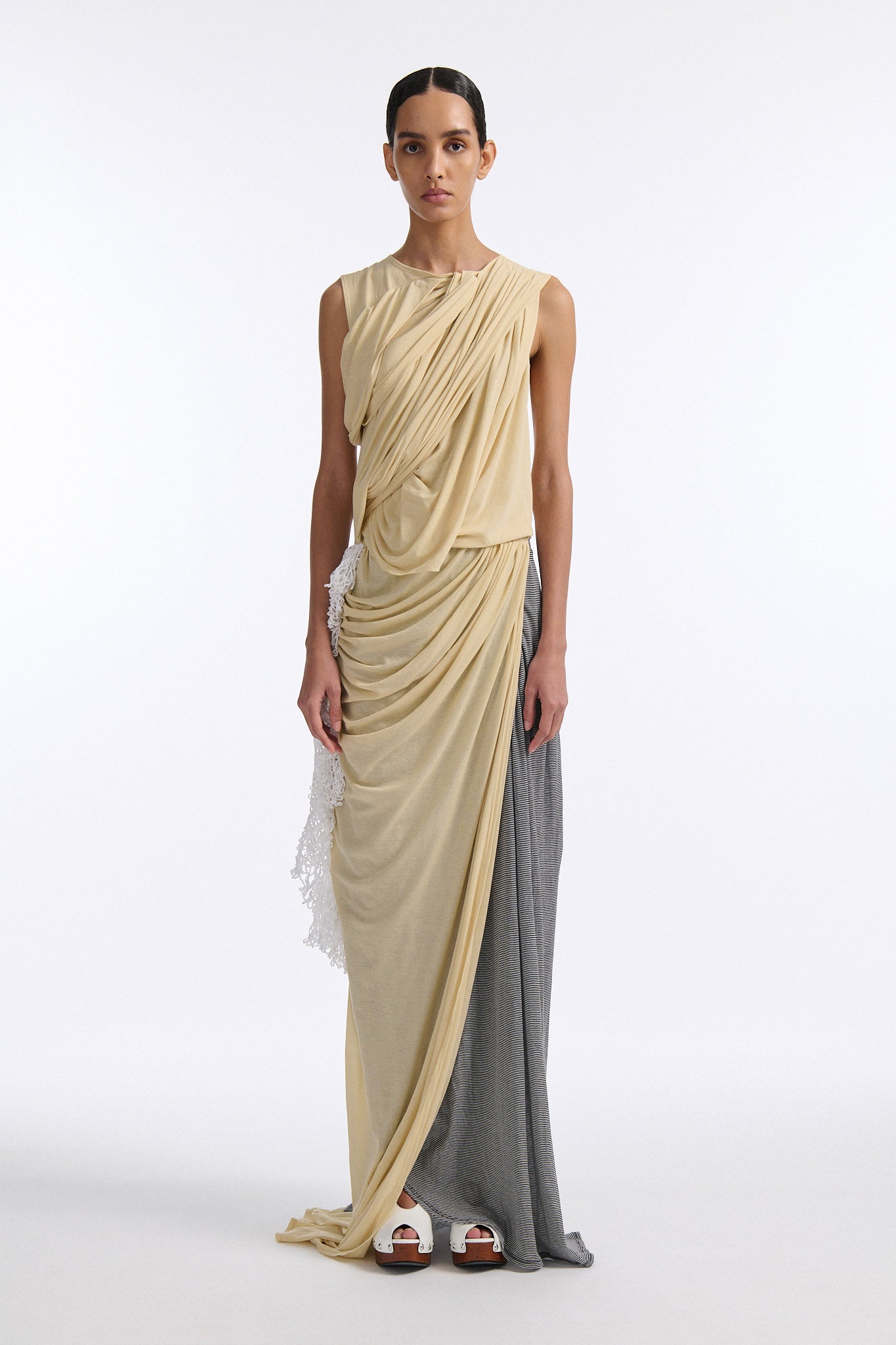 Female model with slicked back black hair in a bun wears fine gauge cotton jersey draped sleeveless gown. Draping goes diagonally across chest and gathers at waist. More diagonal draping on the lower half connects to white crochet at the hip. The front of the dress is beige and the back is horizontal black and white fine stripes.