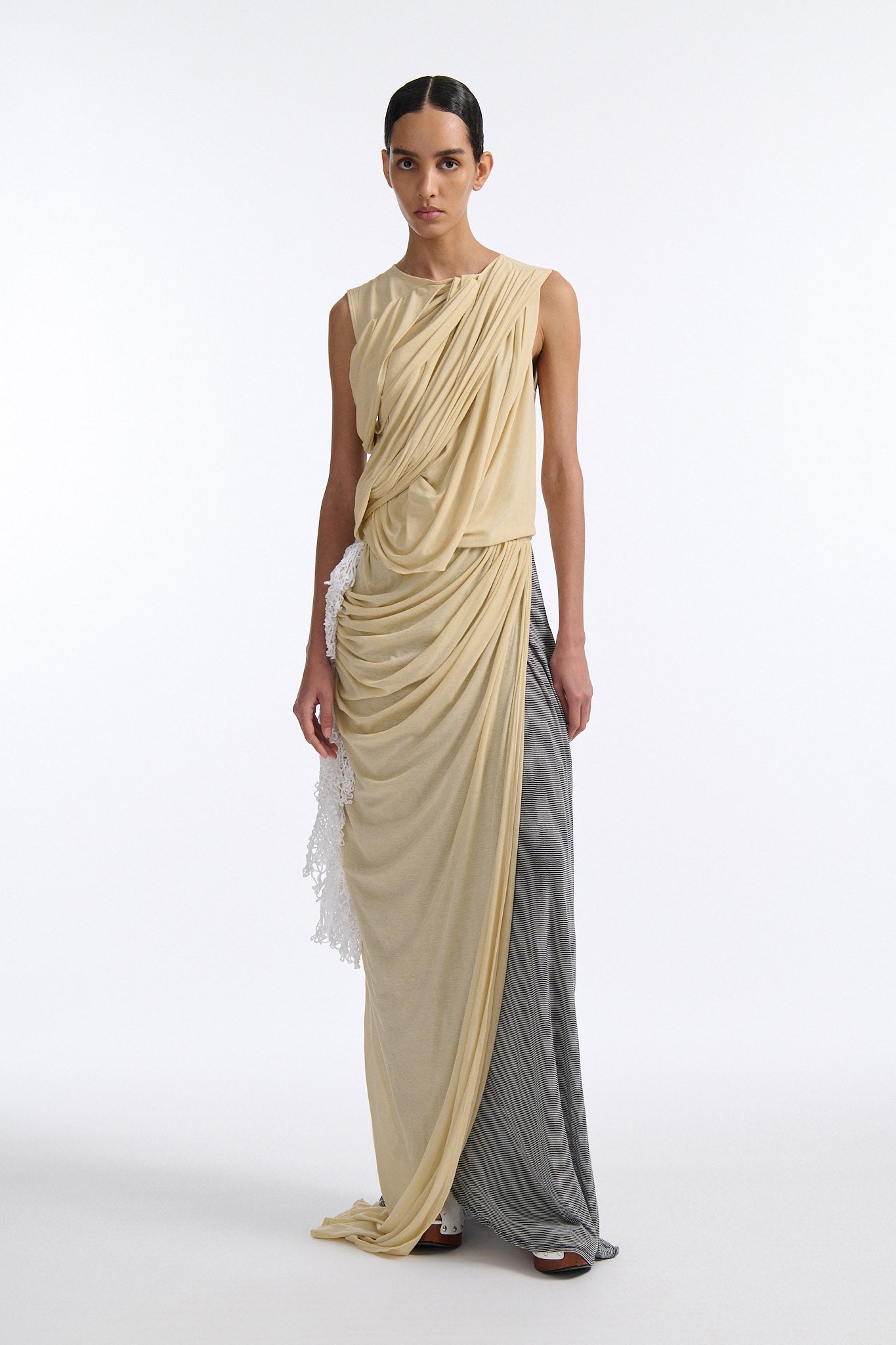 Female model with slicked back black hair in a bun wears fine gauge cotton jersey draped sleeveless gown. Draping goes diagonally across chest and gathers at waist. More diagonal draping on the lower half connects to white crochet at the hip. The front of the dress is beige and the back is horizontal black and white fine stripes.