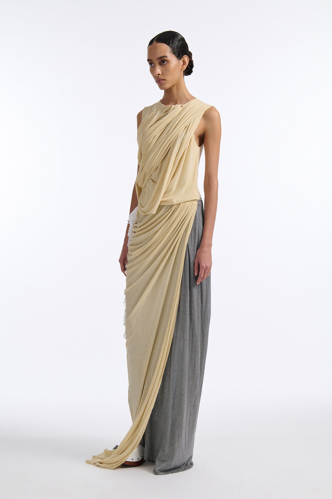 Female model with slicked back black hair in a bun wears fine gauge cotton jersey draped sleeveless gown. Draping goes diagonally across chest and gathers at waist. More diagonal draping on the lower half connects to white crochet at the hip. The front of the dress is beige and the back is horizontal black and white fine stripes.