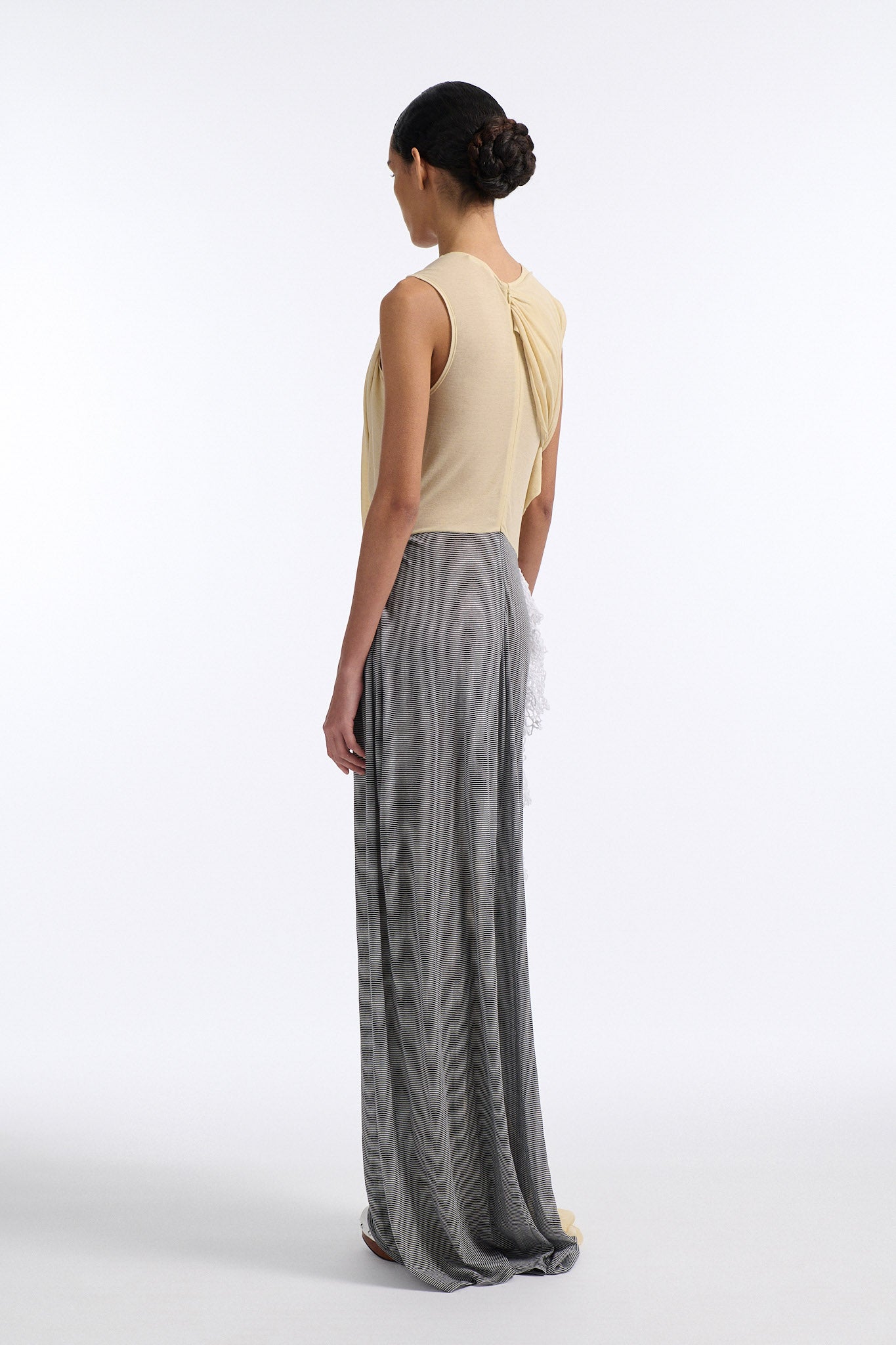 Female model with slicked back black hair in a bun wears fine gauge cotton jersey draped sleeveless gown. Draping goes diagonally across chest and gathers at waist. More diagonal draping on the lower half connects to white crochet at the hip. The front of the dress is beige and the back is horizontal black and white fine stripes.
