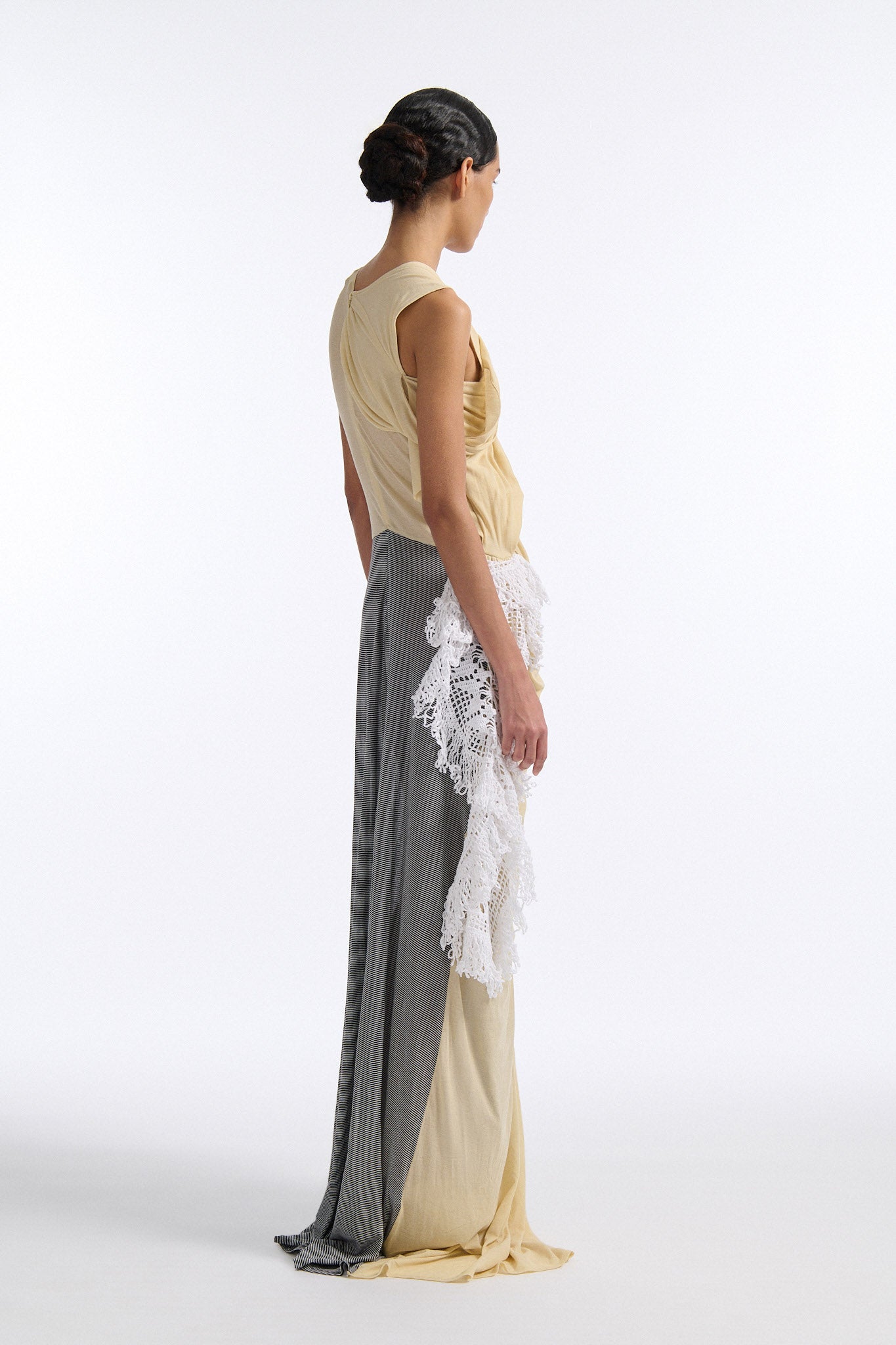 Female model with slicked back black hair in a bun wears fine gauge cotton jersey draped sleeveless gown. Draping goes diagonally across chest and gathers at waist. More diagonal draping on the lower half connects to white crochet at the hip. The front of the dress is beige and the back is horizontal black and white fine stripes.