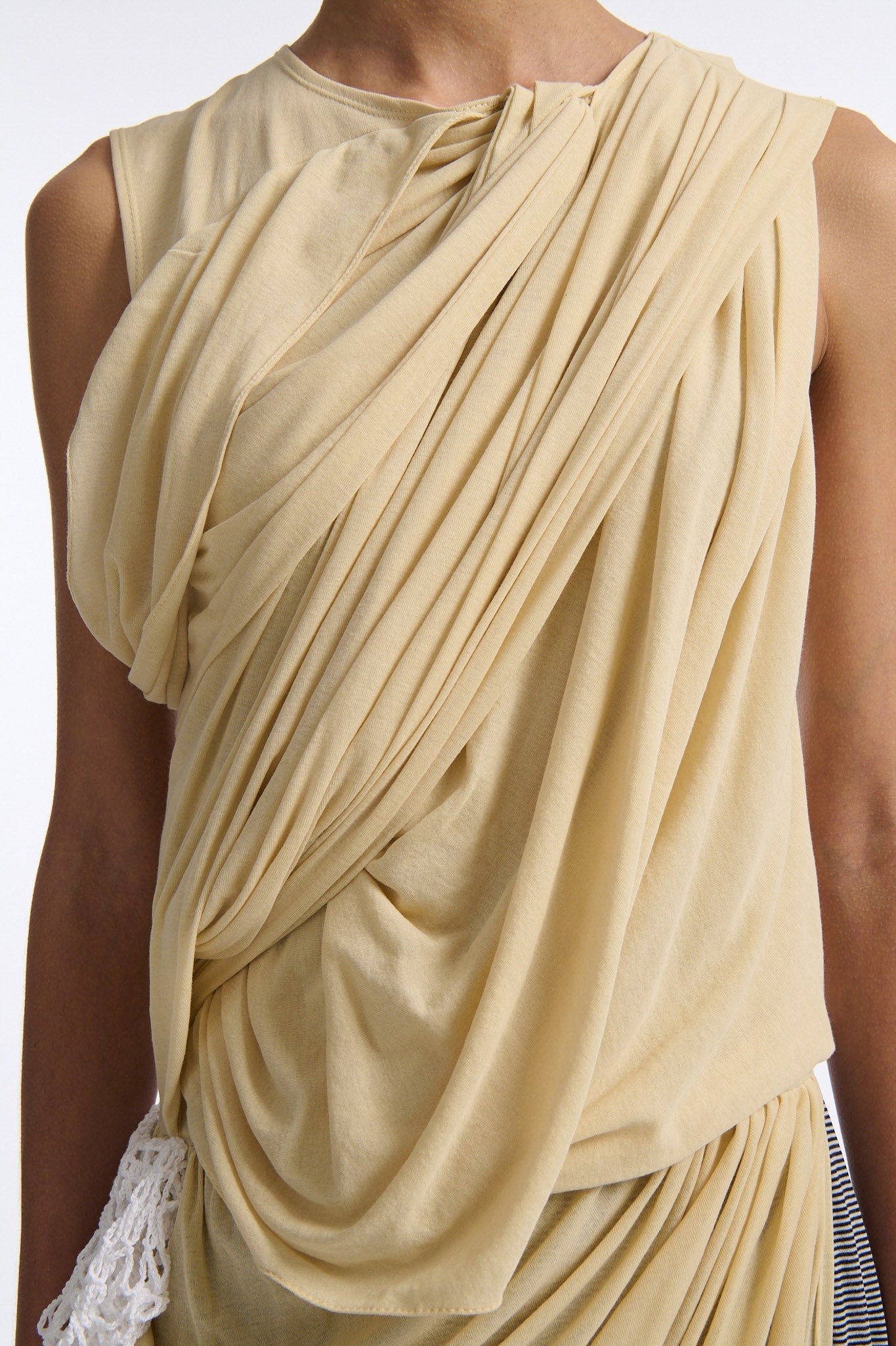 Female model with slicked back black hair in a bun wears fine gauge cotton jersey draped sleeveless gown. Draping goes diagonally across chest and gathers at waist. More diagonal draping on the lower half connects to white crochet at the hip. The front of the dress is beige and the back is horizontal black and white fine stripes.