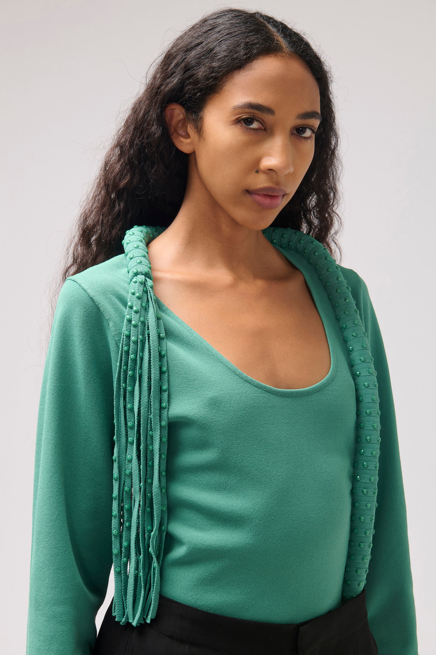 A woman wearing a teal green knit top with a matching fringed cardigan draped over her shoulders, photographed against a neutral light gray studio background.
