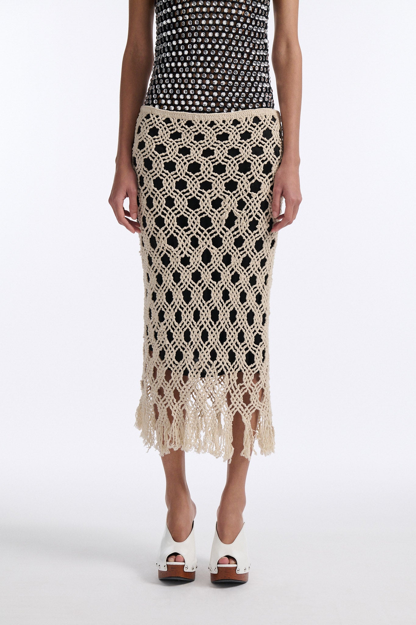 Female model with slicked back black hair in a bun wears black chunky mesh tank top with clear hand-sewn crystals and a cream colored macrame skirt with a black lining.
