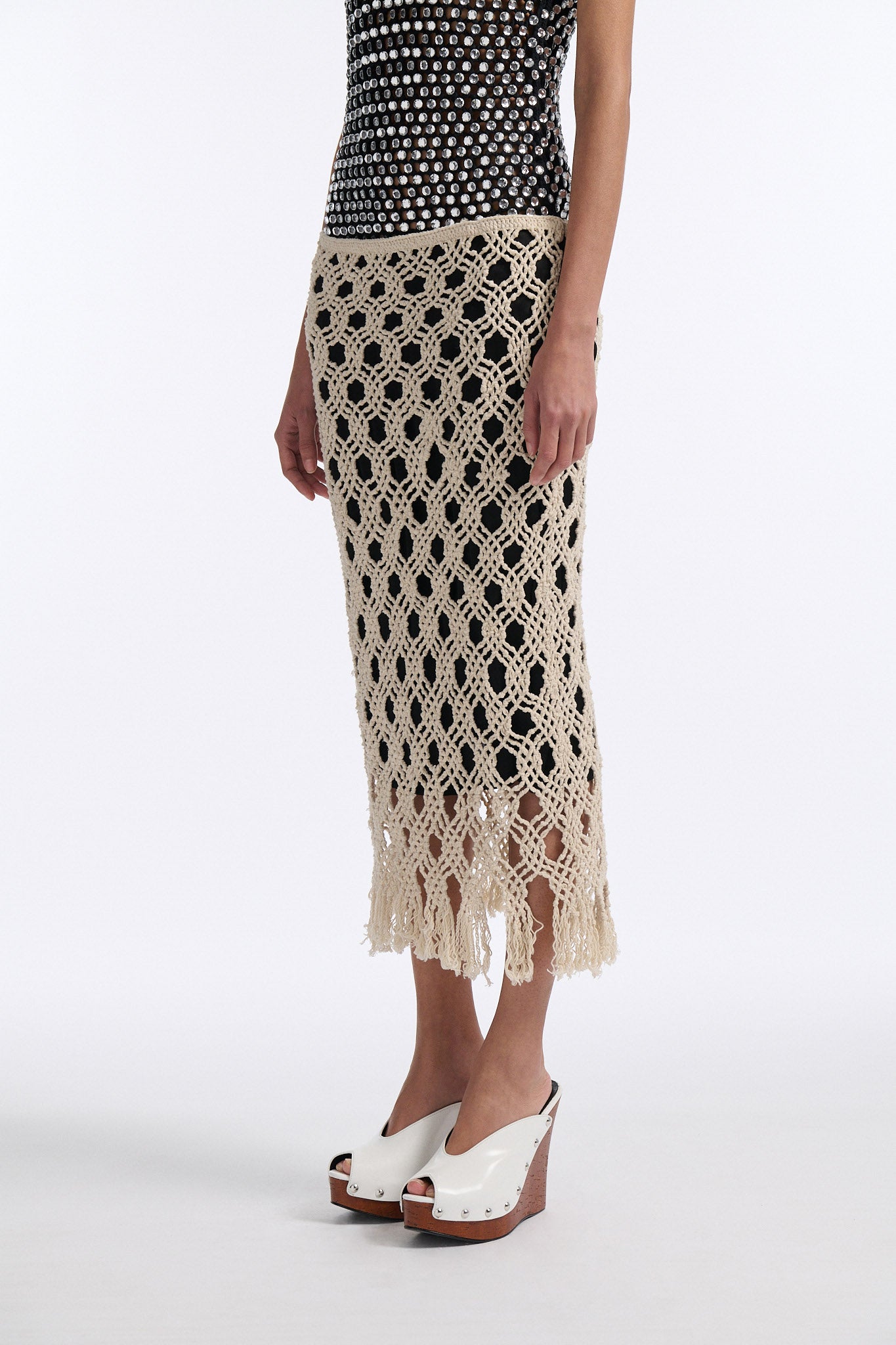 Female model with slicked back black hair in a bun wears black chunky mesh tank top with clear hand-sewn crystals and a cream colored macrame skirt with a black lining.