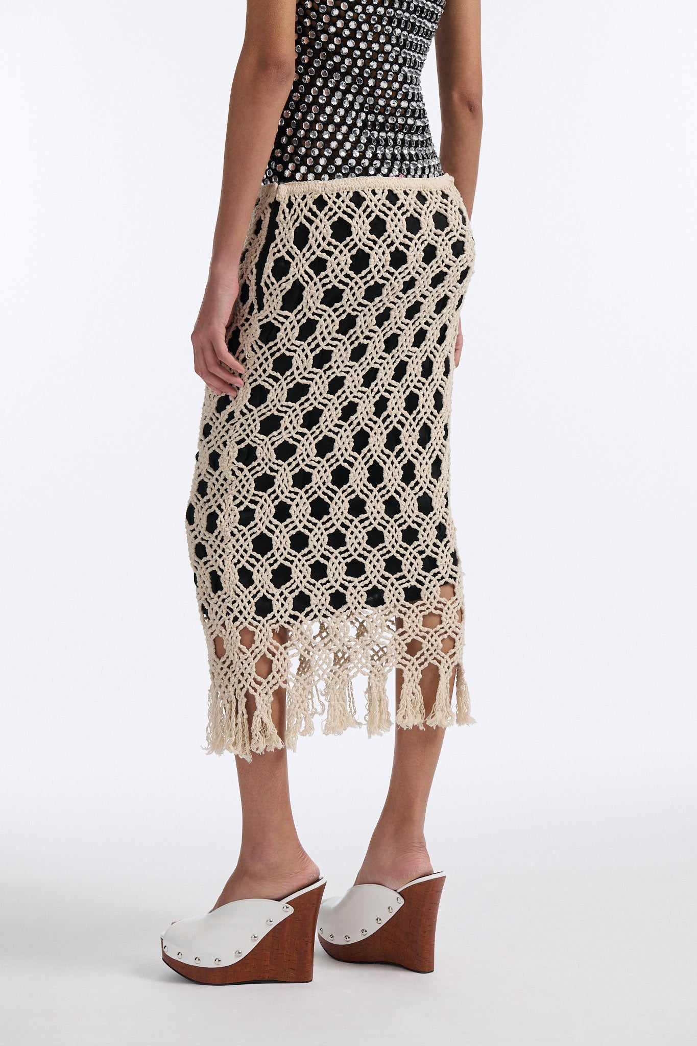 Female model with slicked back black hair in a bun wears black chunky mesh tank top with clear hand-sewn crystals and a cream colored macrame skirt with a black lining.