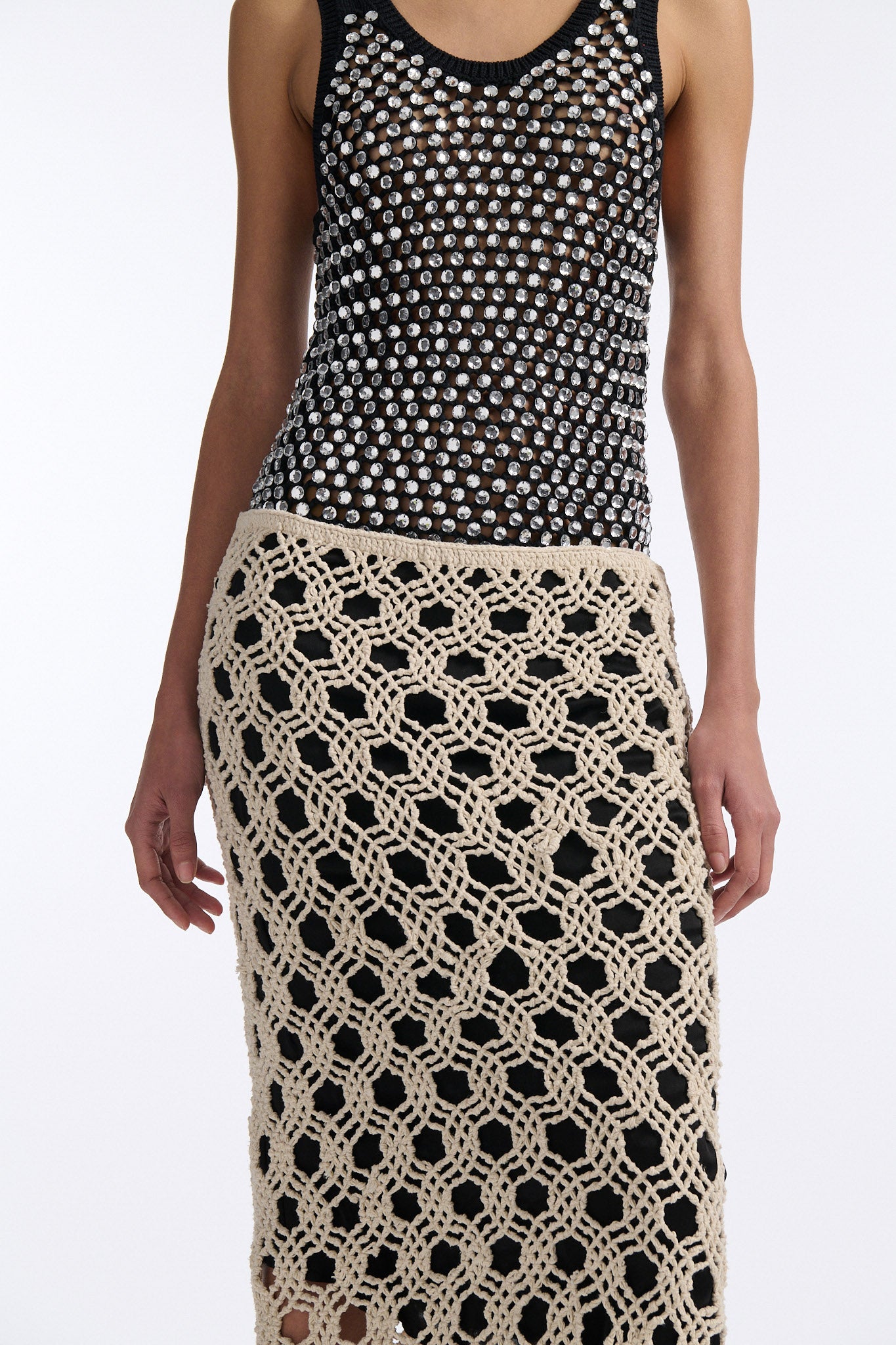 Female model with slicked back black hair in a bun wears black chunky mesh tank top with clear hand-sewn crystals and a cream colored macrame skirt with a black lining.