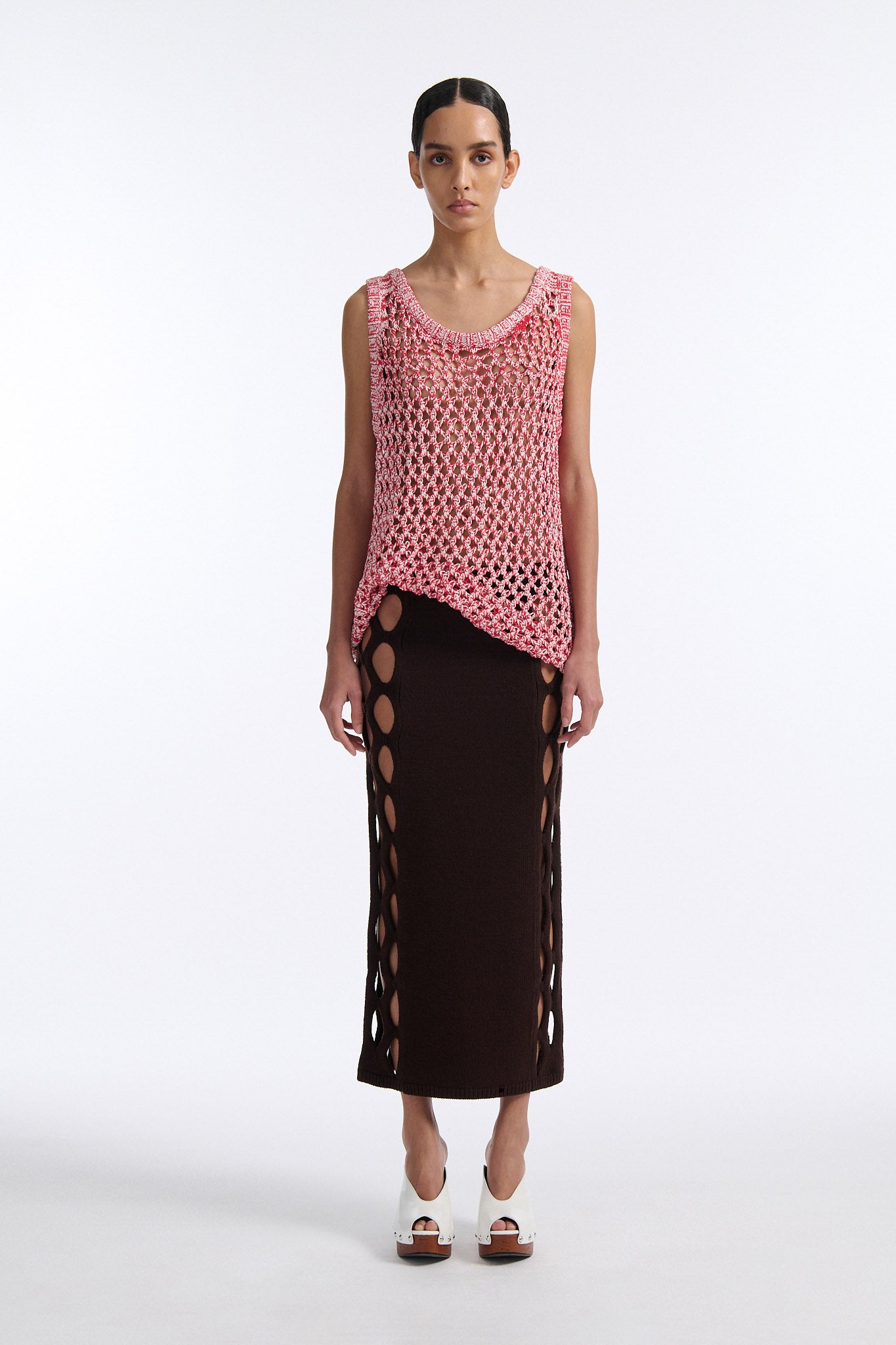 Female model with slicked back black hair in a bun wears white and red mixed stitch chunky mesh see-through tank top. Model also wears brown knit skirt with diamond cutouts on either side.