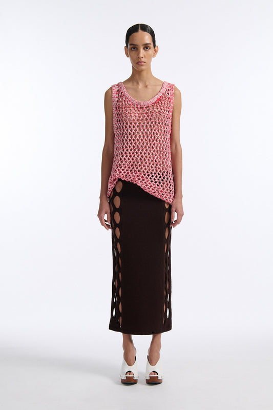 Female model with slicked back black hair in a bun wears white and red mixed stitch chunky mesh see-through tank top. Model also wears brown knit skirt with diamond cutouts on either side.