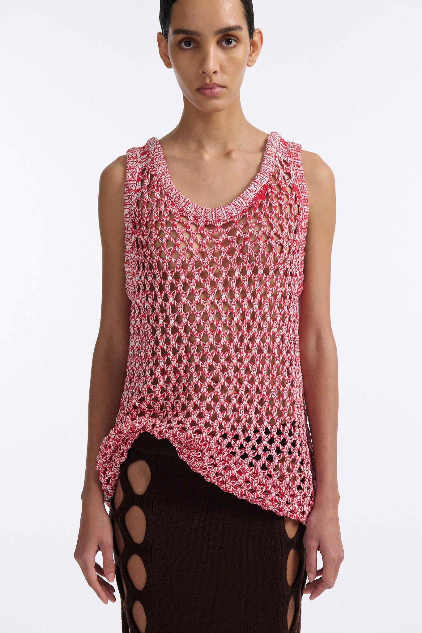 Female model with slicked back black hair in a bun wears white and red mixed stitch chunky mesh see-through tank top. Model also wears brown knit skirt with diamond cutouts on either side.