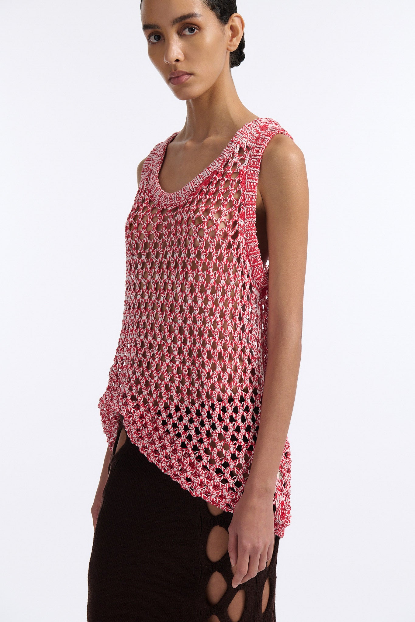 Female model with slicked back black hair in a bun wears white and red mixed stitch chunky mesh see-through tank top. Model also wears brown knit skirt with diamond cutouts on either side.