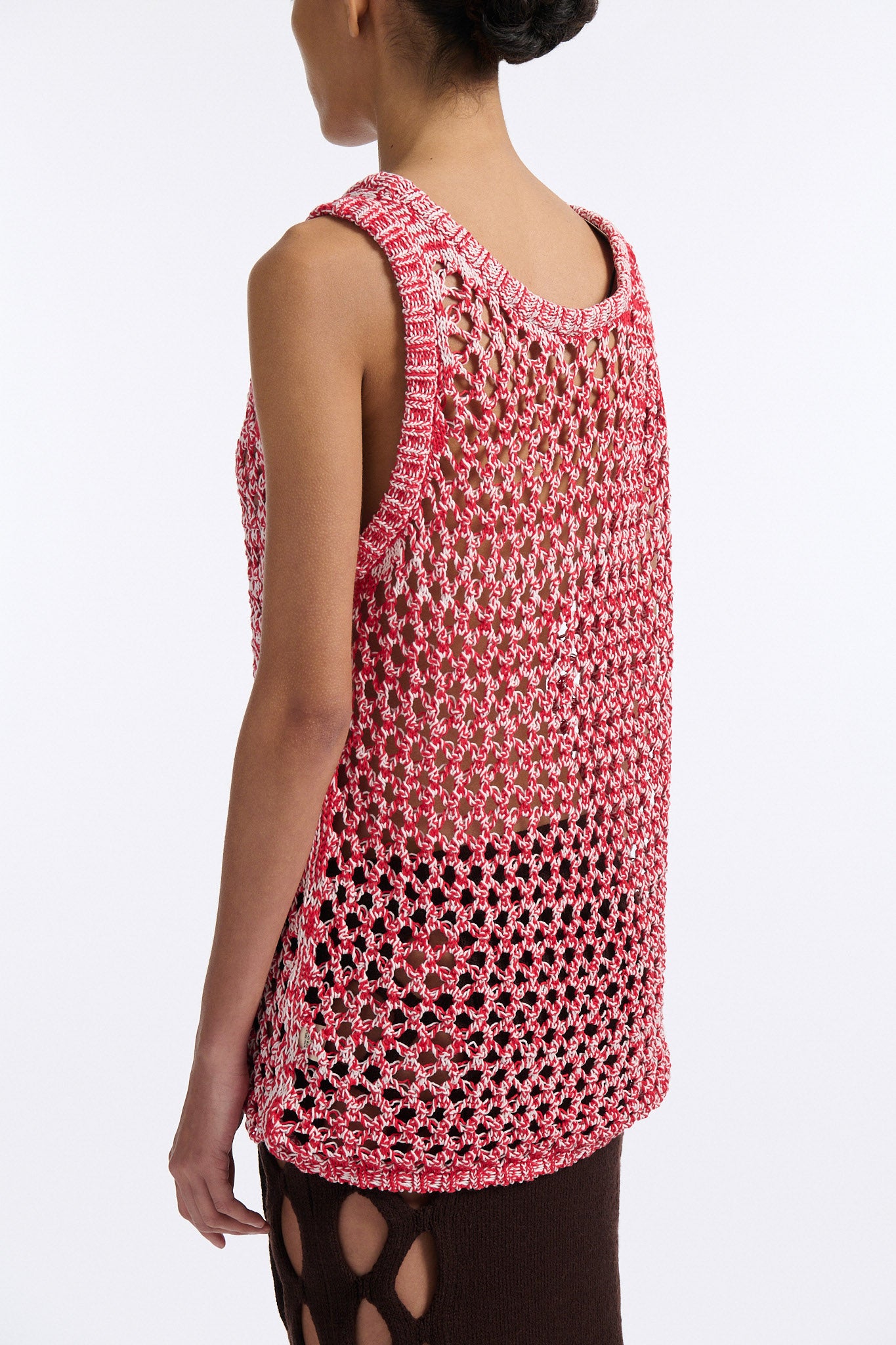 Female model with slicked back black hair in a bun wears white and red mixed stitch chunky mesh see-through tank top. Model also wears brown knit skirt with diamond cutouts on either side.