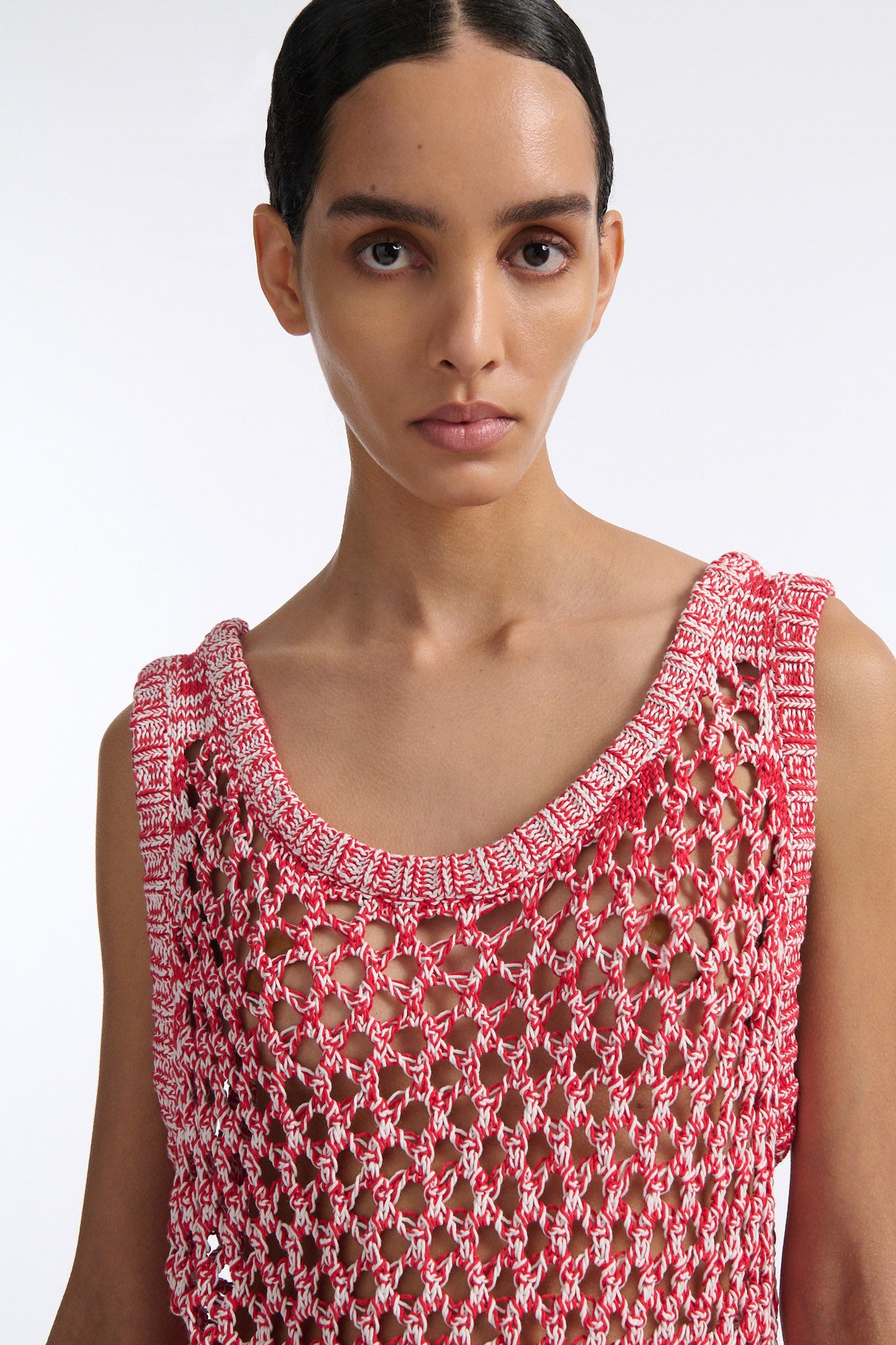 Female model with slicked back black hair in a bun wears white and red mixed stitch chunky mesh see-through tank top. Model also wears brown knit skirt with diamond cutouts on either side.