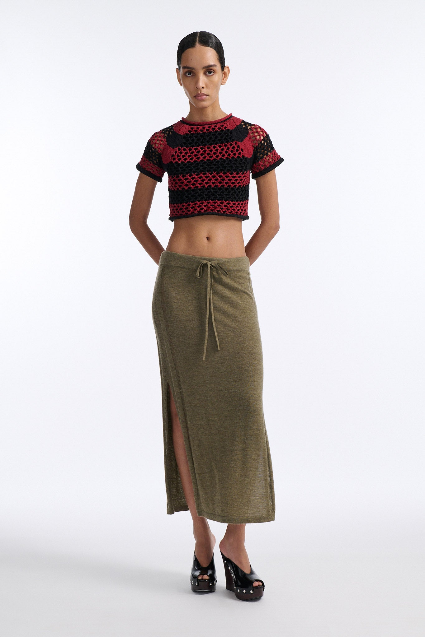 Female model with slicked back black hair in a bun wears chunky mesh horizontal striped cropped tee in red and black. The model also wears a fine gauge drawstring cashmere skirt with pointelle stitching on either side and a long slit on either side.