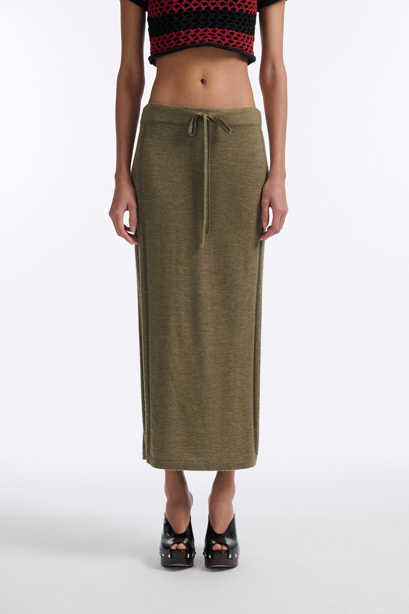 Female model with slicked back black hair in a bun wears chunky mesh horizontal striped cropped tee in red and black. The model also wears a fine gauge drawstring cashmere skirt with pointelle stitching on either side and a long slit on either side.