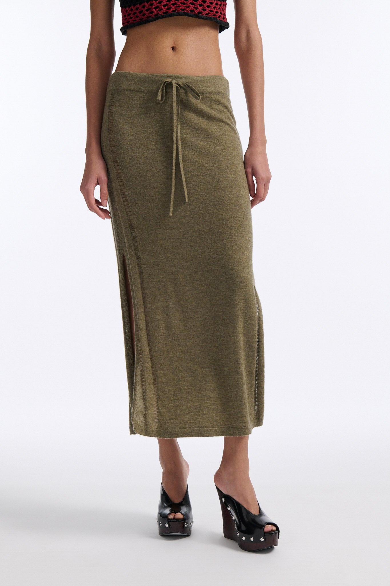 Female model with slicked back black hair in a bun wears chunky mesh horizontal striped cropped tee in red and black. The model also wears a fine gauge drawstring cashmere skirt with pointelle stitching on either side and a long slit on either side.