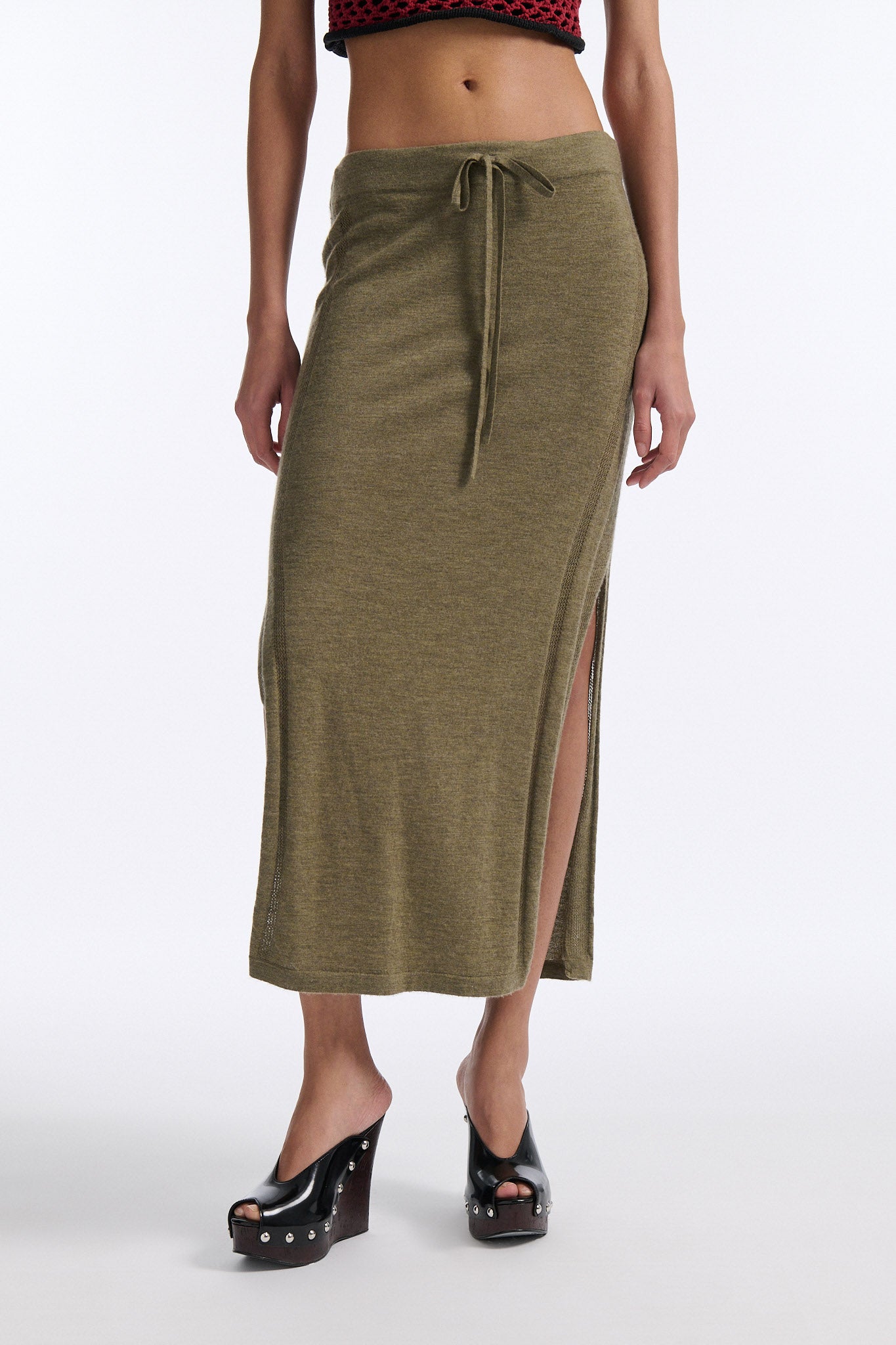 Female model with slicked back black hair in a bun wears chunky mesh horizontal striped cropped tee in red and black. The model also wears a fine gauge drawstring cashmere skirt with pointelle stitching on either side and a long slit on either side.