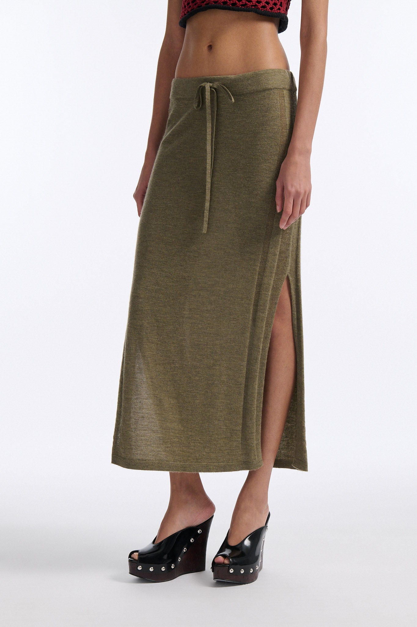 Female model with slicked back black hair in a bun wears chunky mesh horizontal striped cropped tee in red and black. The model also wears a fine gauge drawstring cashmere skirt with pointelle stitching on either side and a long slit on either side.