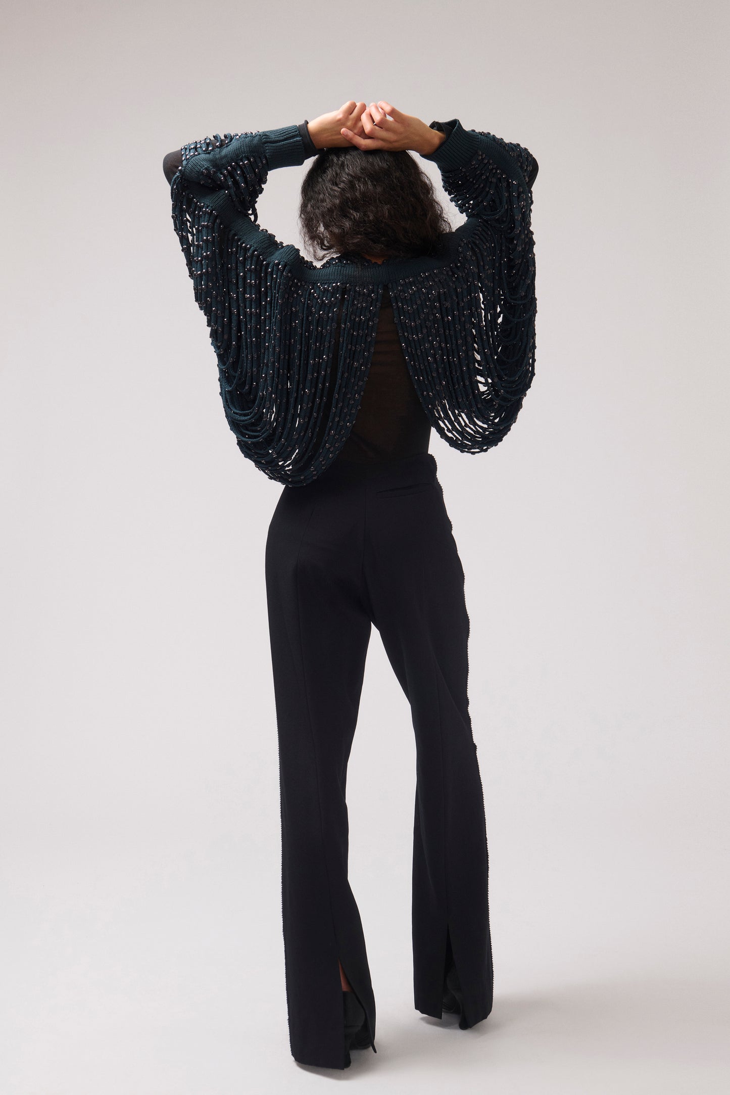 A model viewed from behind wearing a dark teal beaded knit cardigan with dramatic draped sleeves and black flared pants, set against a neutral light gray studio background.