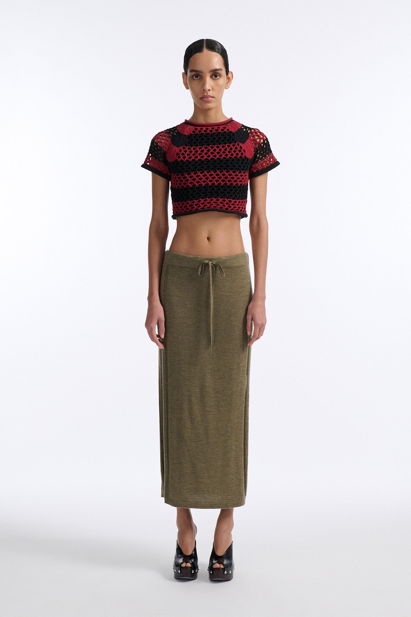 Female model with slicked back black hair in a bun wears red and black bold striped mesh tee, cropped above the belly button, and mid-waist long adjustable skirt in soft green cashmere with black platform wedges.