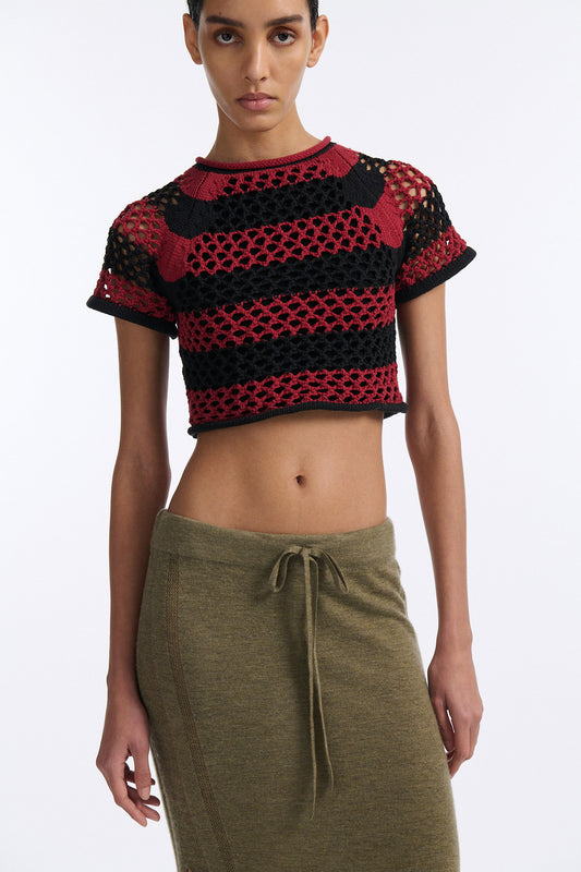 Female model with slicked back black hair in a bun wears red and black bold striped mesh tee, cropped above the belly button, and mid-waist long adjustable skirt in soft green cashmere with black platform wedges.