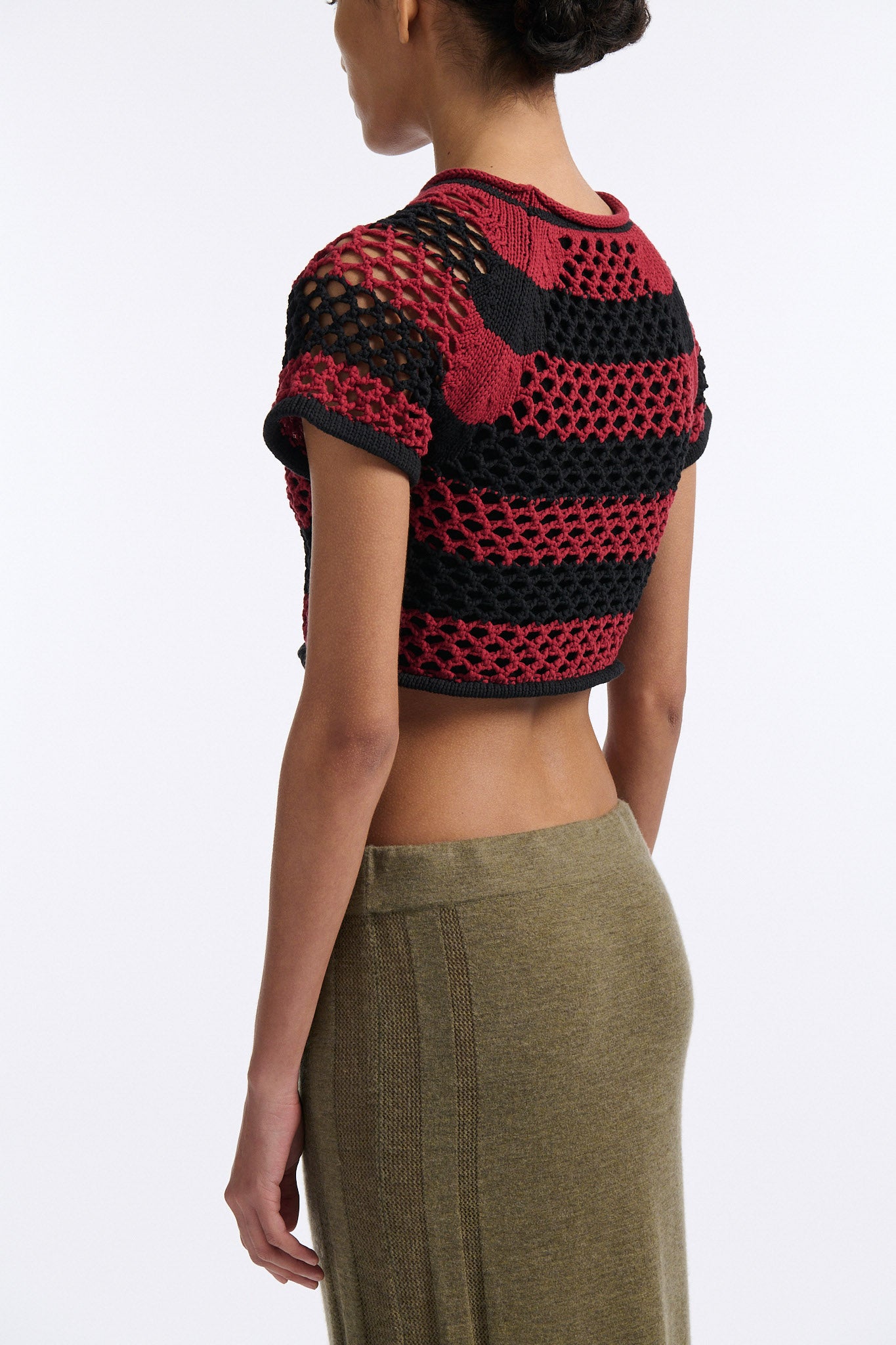 Female model with slicked back black hair in a bun wears red and black bold striped mesh tee, cropped above the belly button, and mid-waist long adjustable skirt in soft green cashmere with black platform wedges.