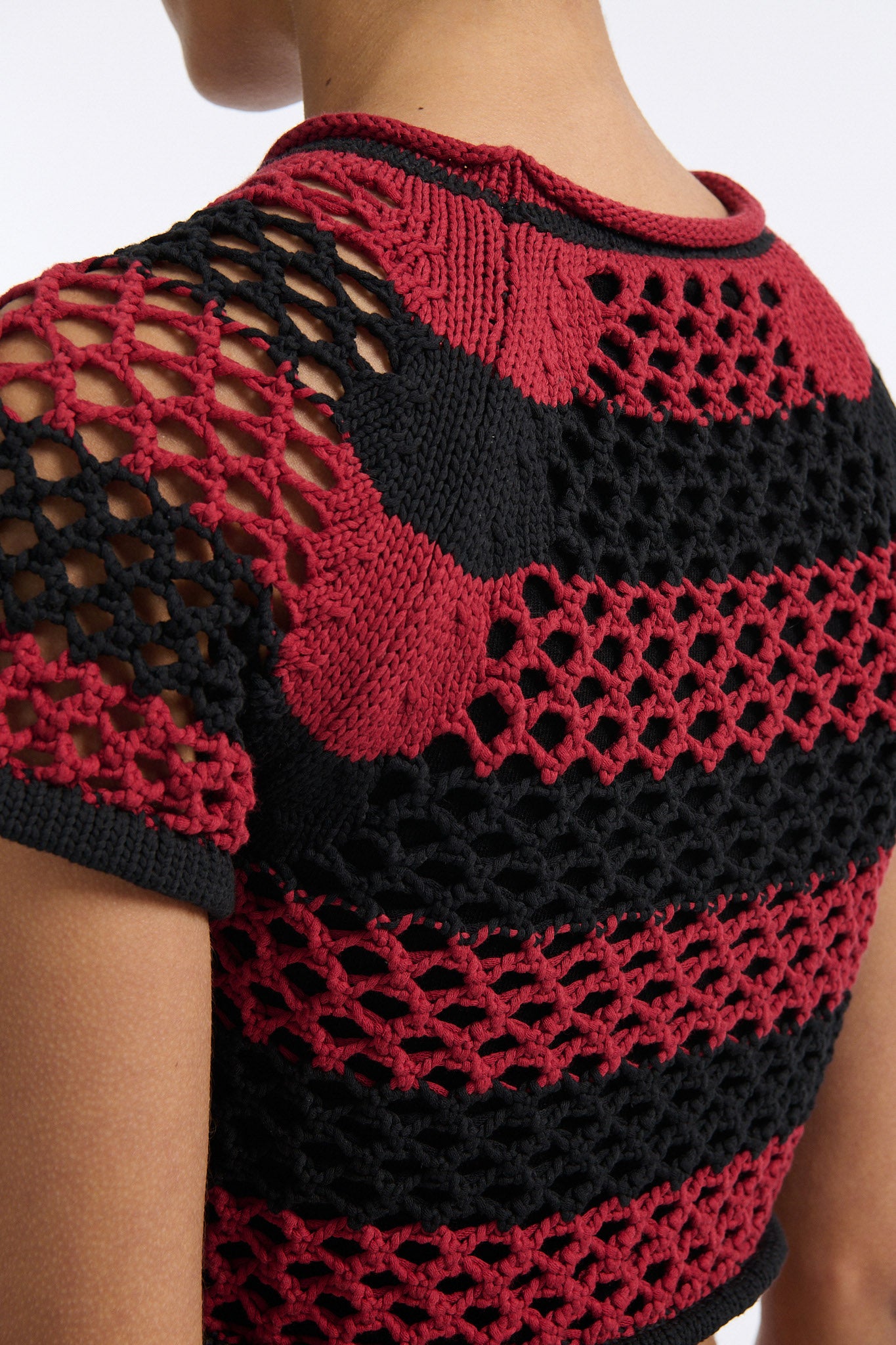 Female model with slicked back black hair in a bun wears red and black bold striped mesh tee, cropped above the belly button, and mid-waist long adjustable skirt in soft green cashmere with black platform wedges.