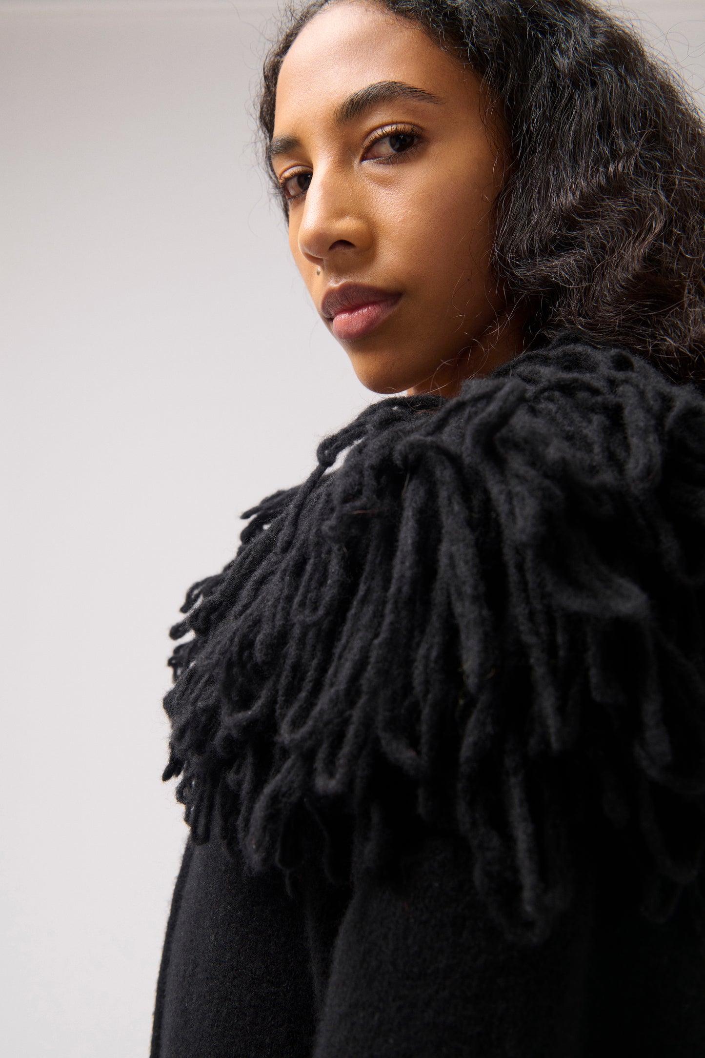 A close-up portrait of a model wearing a black textured knit cardigan with a large, fringed, tassel-like collar against a plain light gray background.