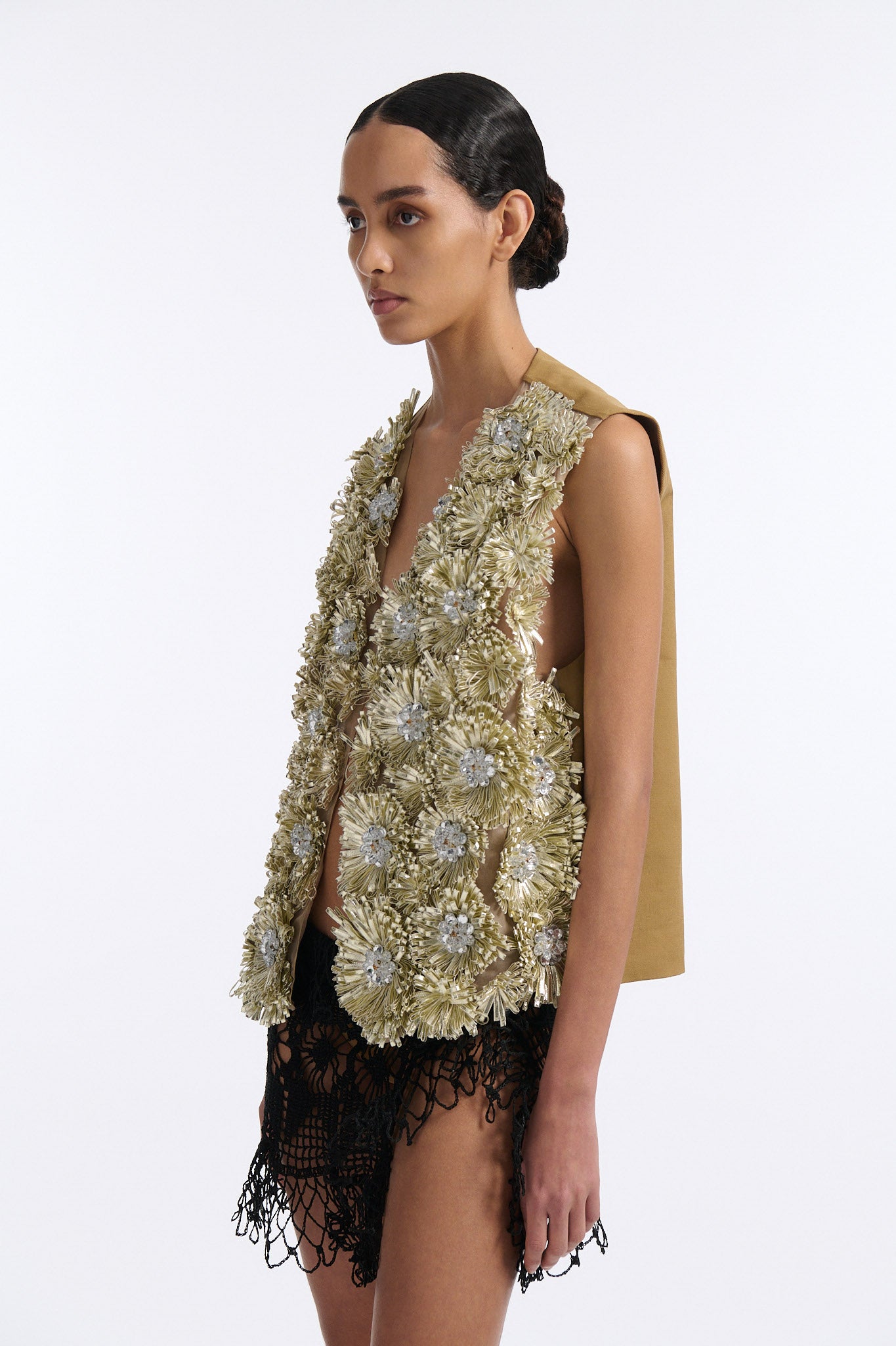 Female model with slicked back black hair in a bun wears metallic ribbon flower V-neck one button vest in pale gold and silver with black see-through crochet mini skirt.