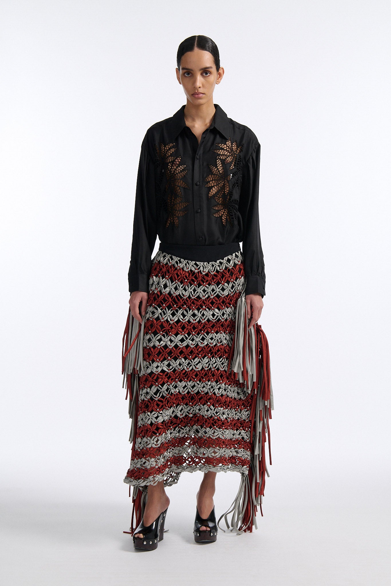 Female model with slicked back black hair in a bun wears black silk button down with palm leaf broderie anglaise on chest and back. Model also wears burnt orange and blue-grey horizontal striped macrame skirt with asymmetric fringe on both sides.