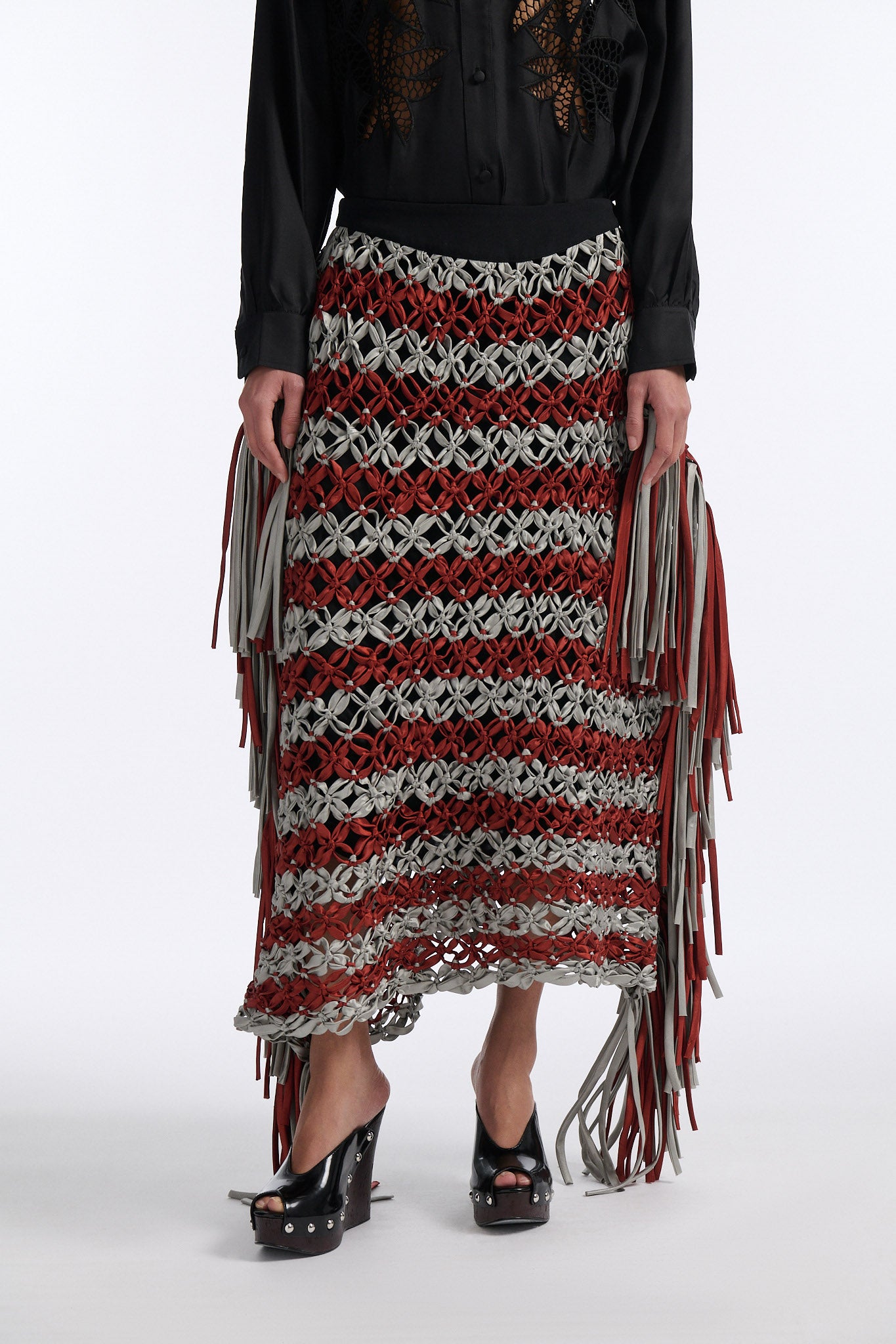 Female model with slicked back black hair in a bun wears black silk button down with palm leaf broderie anglaise on chest and back. Model also wears burnt orange and blue-grey horizontal striped macrame skirt with asymmetric fringe on both sides.