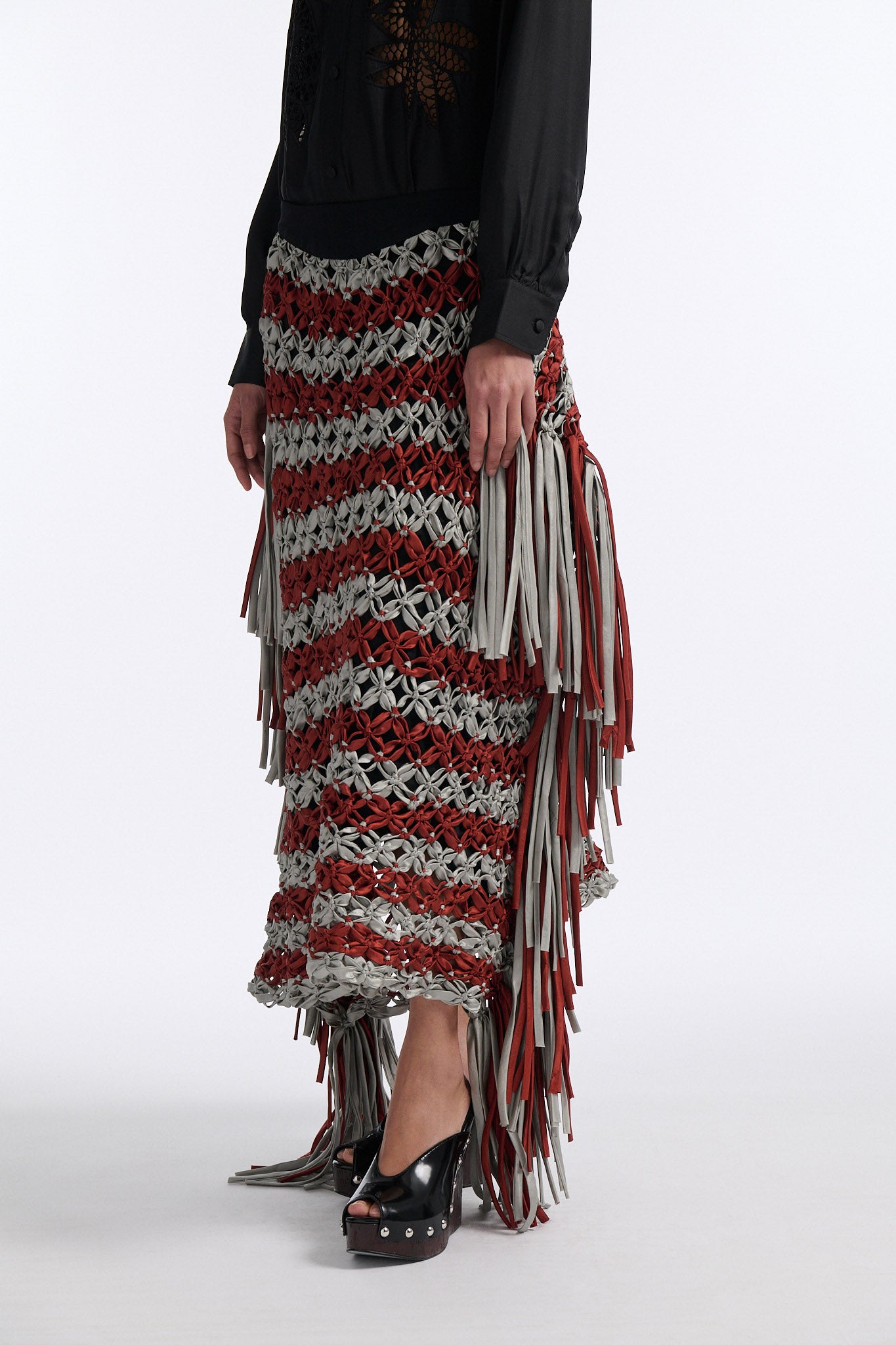 Female model with slicked back black hair in a bun wears black silk button down with palm leaf broderie anglaise on chest and back. Model also wears burnt orange and blue-grey horizontal striped macrame skirt with asymmetric fringe on both sides.