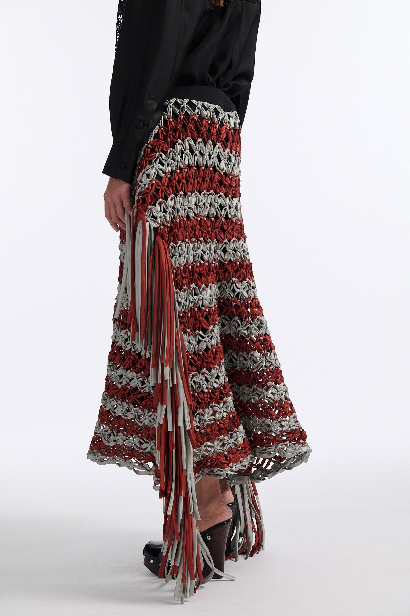 Female model with slicked back black hair in a bun wears black silk button down with palm leaf broderie anglaise on chest and back. Model also wears burnt orange and blue-grey horizontal striped macrame skirt with asymmetric fringe on both sides.