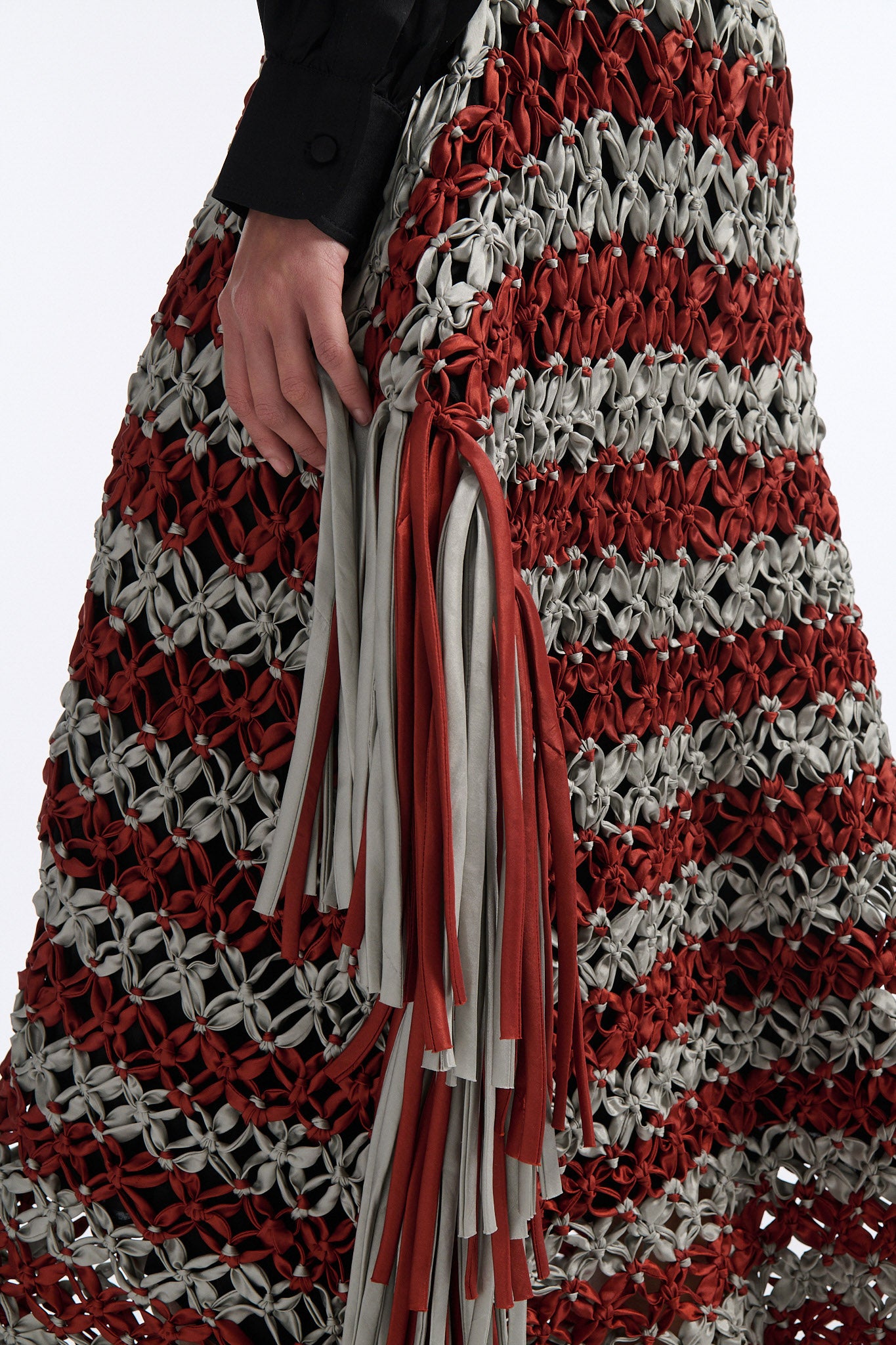 Female model with slicked back black hair in a bun wears black silk button down with palm leaf broderie anglaise on chest and back. Model also wears burnt orange and blue-grey horizontal striped macrame skirt with asymmetric fringe on both sides.