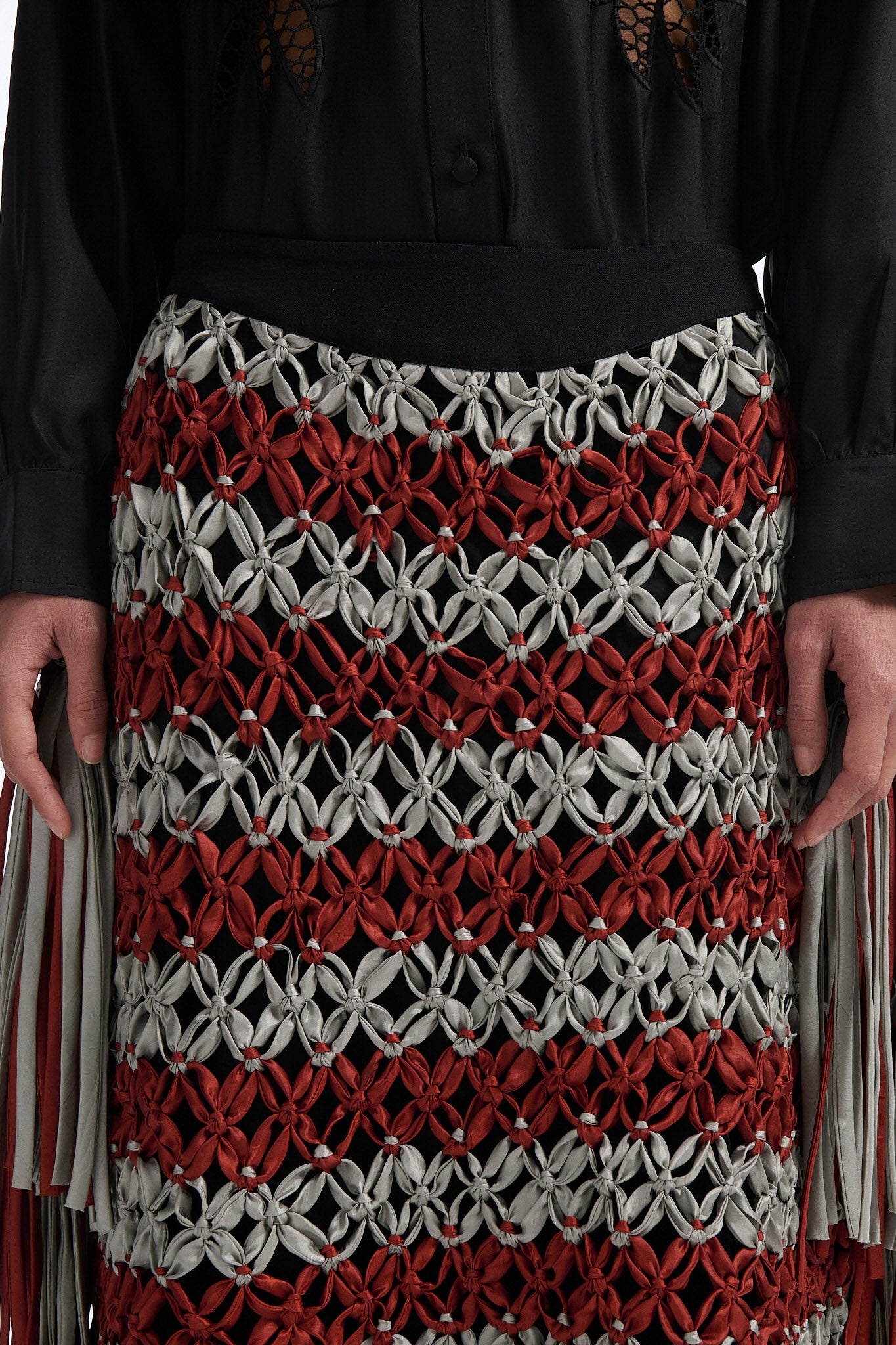 Female model with slicked back black hair in a bun wears black silk button down with palm leaf broderie anglaise on chest and back. Model also wears burnt orange and blue-grey horizontal striped macrame skirt with asymmetric fringe on both sides.