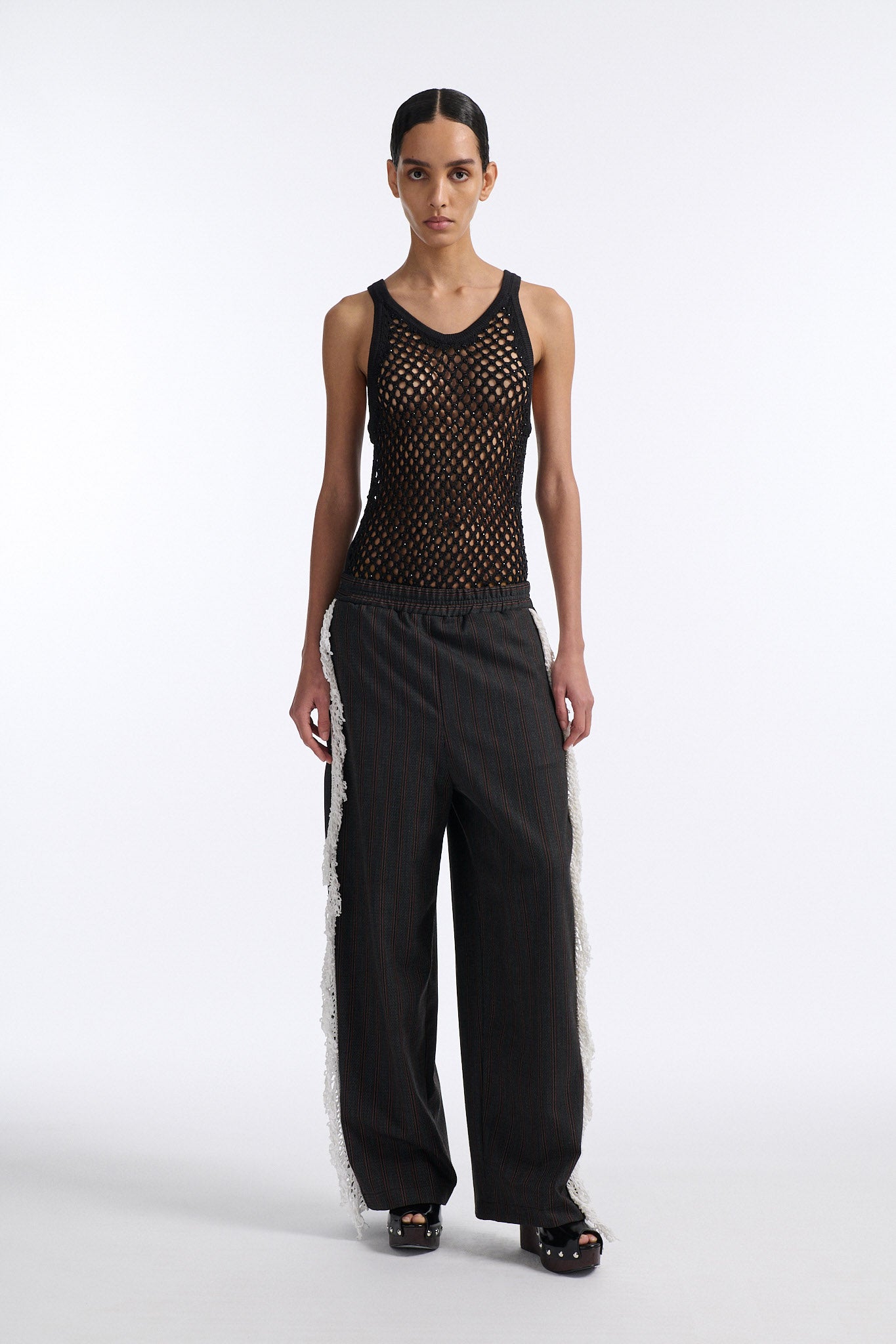Female model with slicked back black hair in a bun wears back fine crystal embellished mesh tank with charcoal grey and burgundy vertical striped trousers. Trousers have elastic waist with white crochet fringe along either leg.