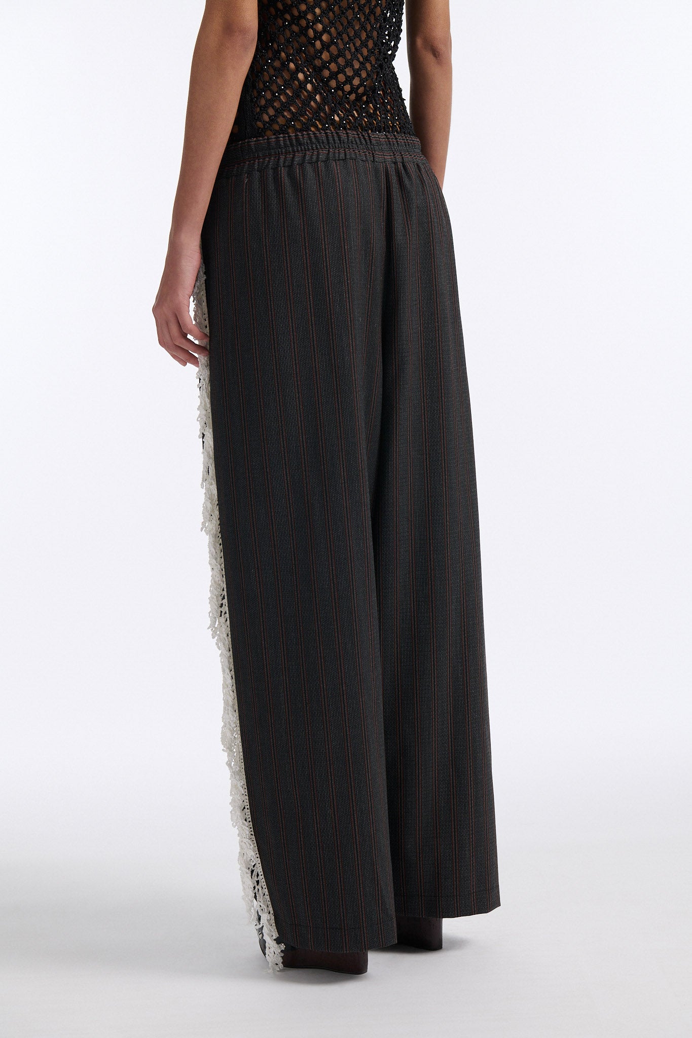 Female model with slicked back black hair in a bun wears back fine crystal embellished mesh tank with charcoal grey and burgundy vertical striped trousers. Trousers have elastic waist with white crochet fringe along either leg.
