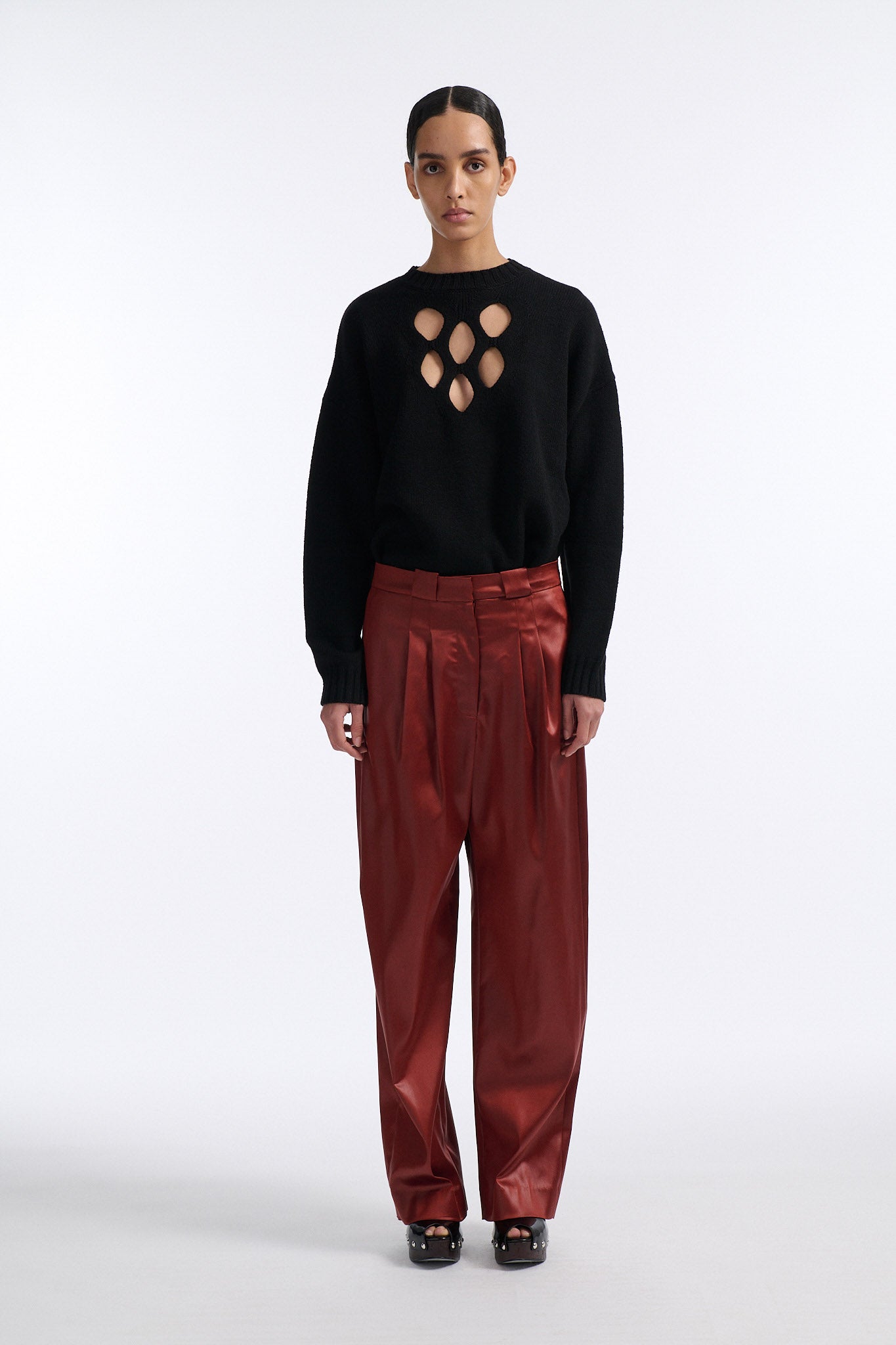 Female model with slicked back black hair in a bun wears black crewneck long sleeve sweater with 5 hole cutout at center of chest. Model also wears shiny rust red pleated trousers with black wedges.