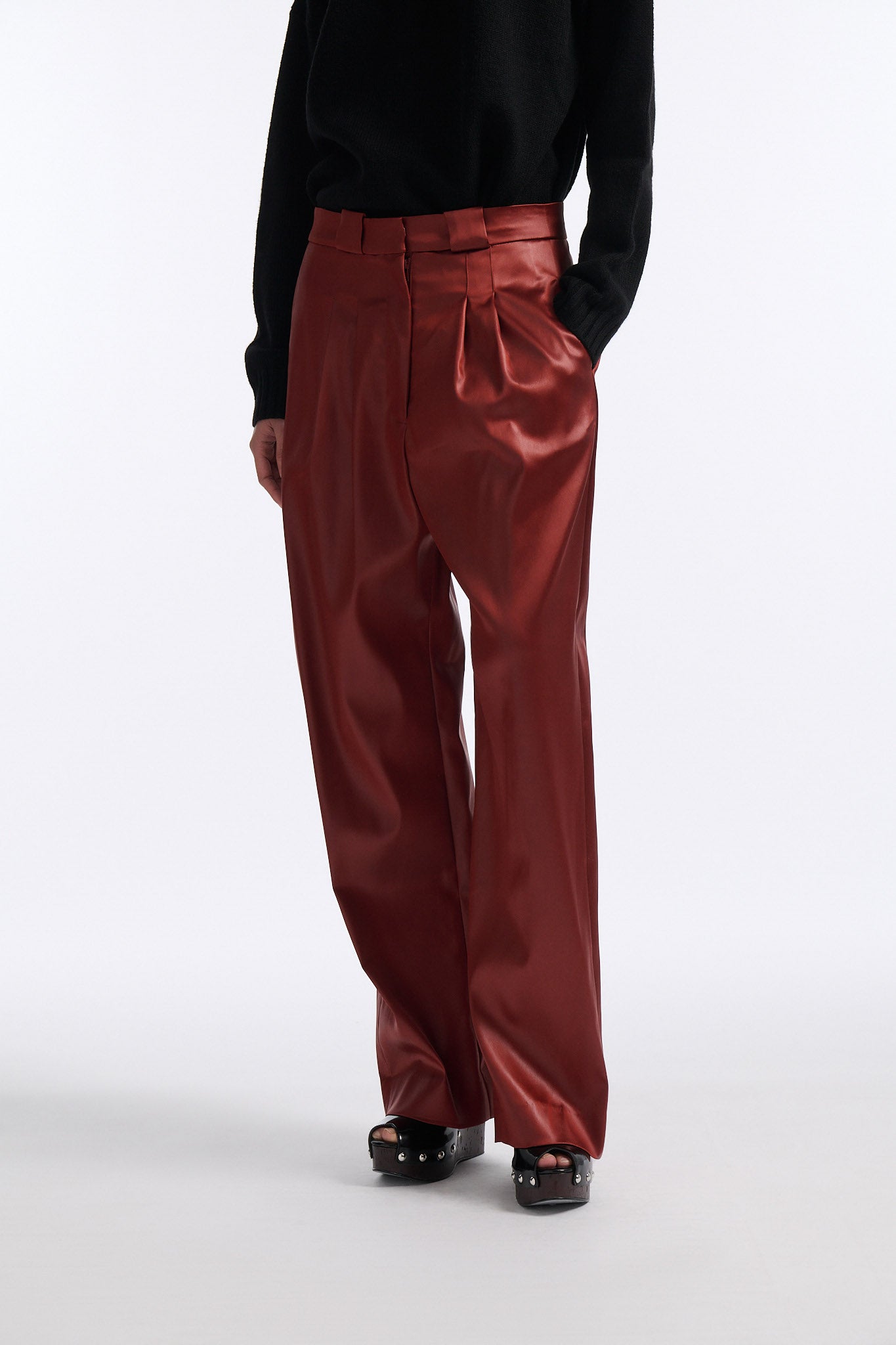 Female model with slicked back black hair in a bun wears black crewneck long sleeve sweater with 5 hole cutout at center of chest. Model also wears shiny rust red pleated trousers with black wedges.