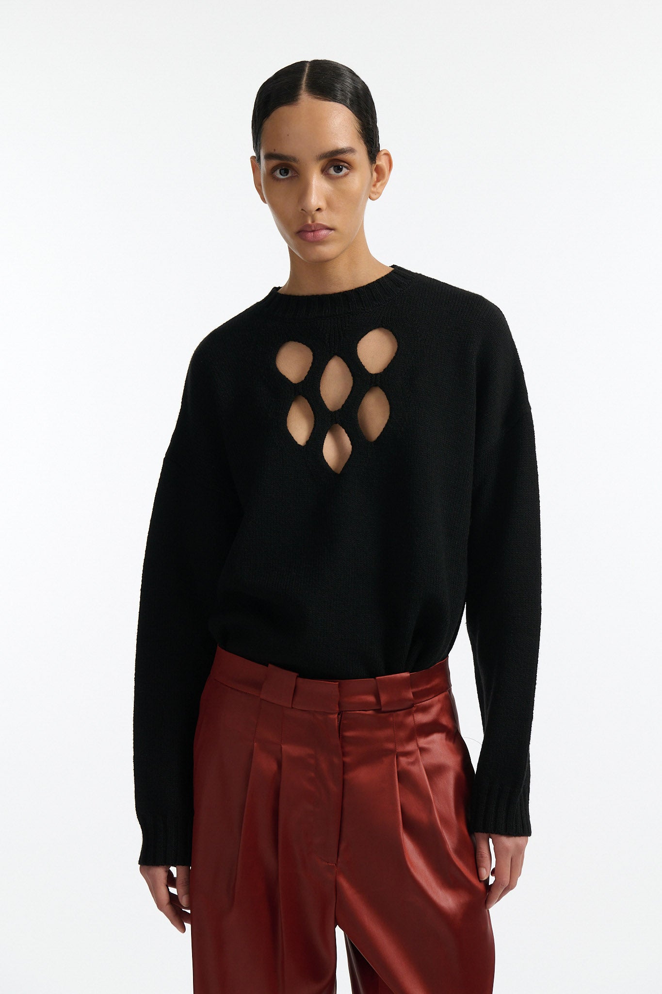 Female model with slicked back black hair in a bun wears black crewneck long sleeve sweater with 5 hole cutout at center of chest. Model also wears shiny rust red pleated trousers with black wedges.
