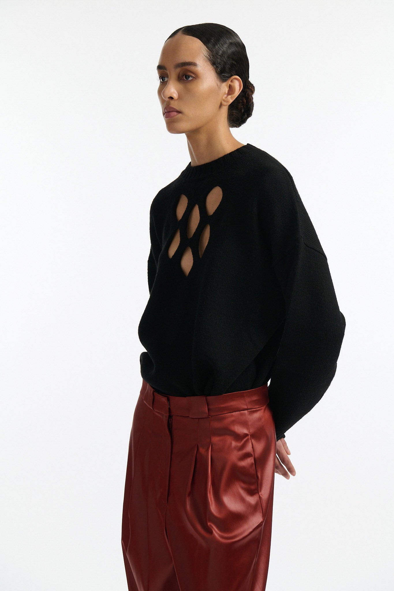Female model with slicked back black hair in a bun wears black crewneck long sleeve sweater with 5 hole cutout at center of chest. Model also wears shiny rust red pleated trousers with black wedges.