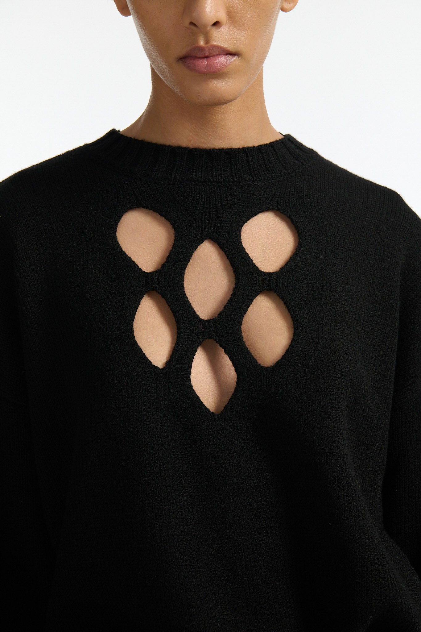 Female model with slicked back black hair in a bun wears black crewneck long sleeve sweater with 5 hole cutout at center of chest. Model also wears shiny rust red pleated trousers with black wedges.