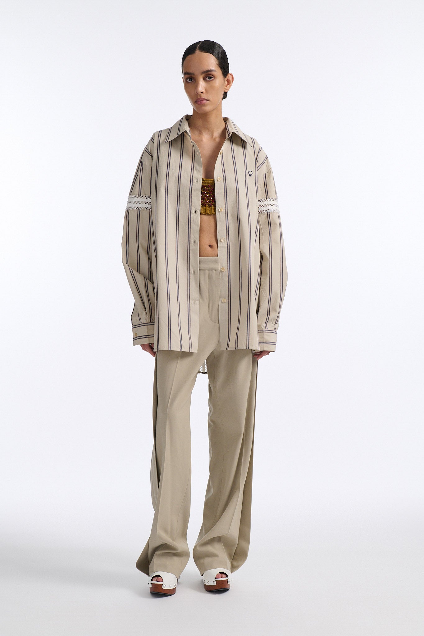Female model with slicked back black hair in a bun wears vertical striped ecru button-down shirt with hand stitched stripes around either upper-arm. Model also wears ecru pleated trouser with extra panel on sides for volume.