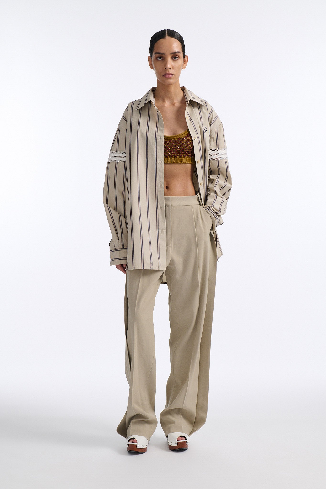 Female model with slicked back black hair in a bun wears vertical striped ecru button-down shirt with hand stitched stripes around either upper-arm. Model also wears ecru pleated trouser with extra panel on sides for volume.
