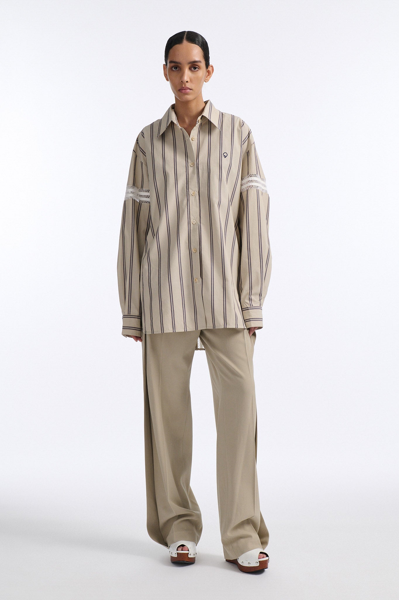 Female model with slicked back black hair in a bun wears vertical striped ecru button-down shirt with hand stitched stripes around either upper-arm. Model also wears ecru pleated trouser with extra panel on sides for volume.