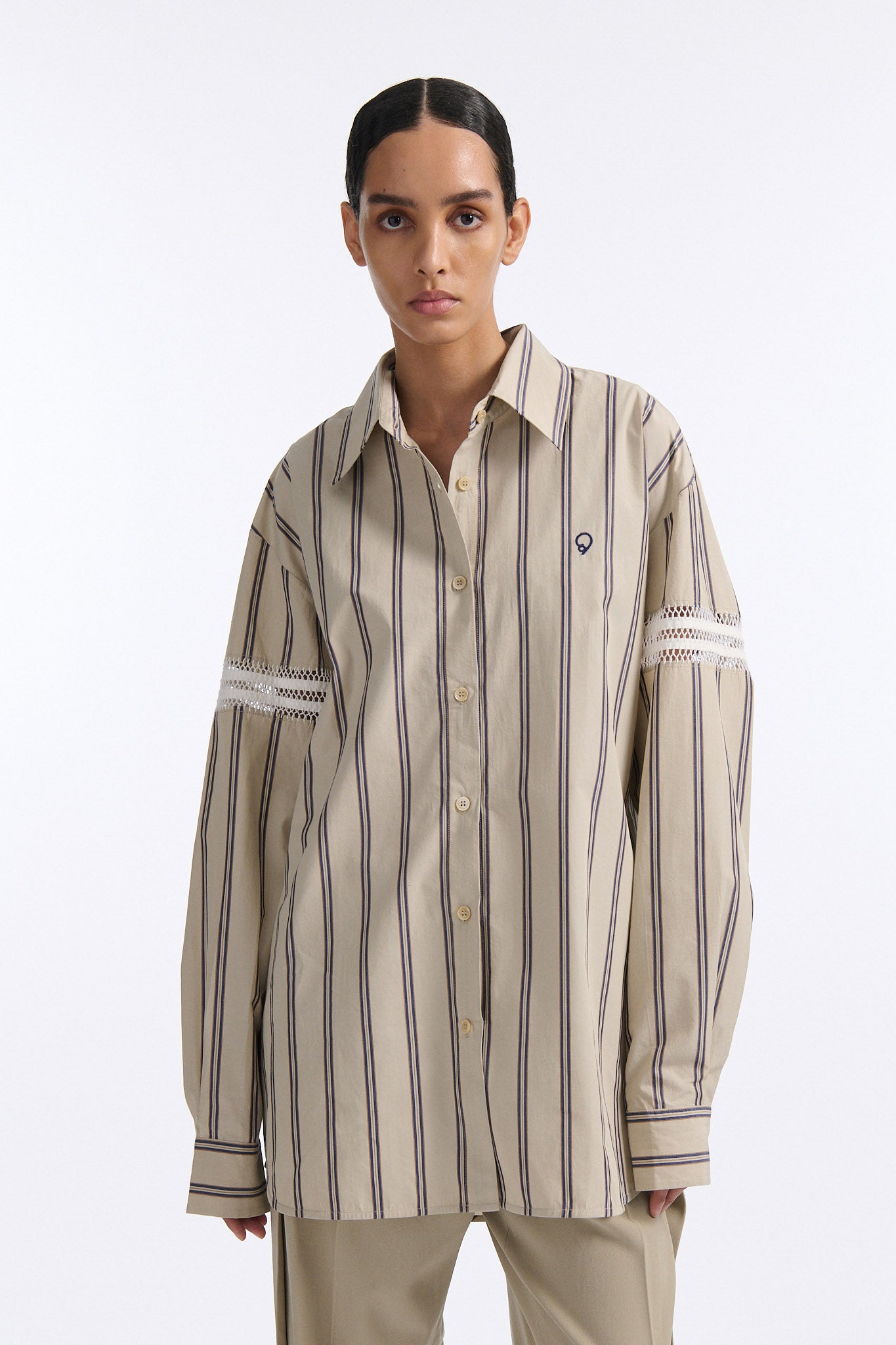 Female model with slicked back black hair in a bun wears vertical striped ecru button-down shirt with hand stitched stripes around either upper-arm. Model also wears ecru pleated trouser with extra panel on sides for volume.