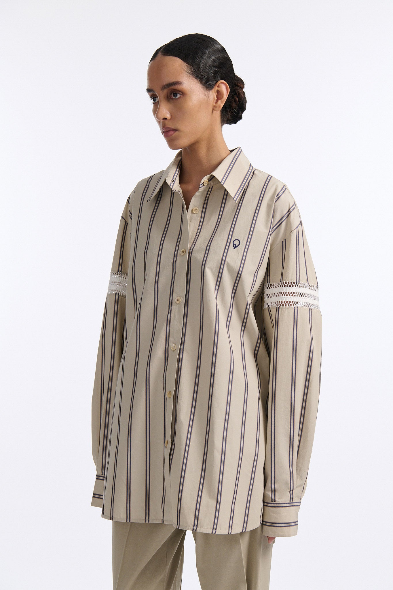 Female model with slicked back black hair in a bun wears vertical striped ecru button-down shirt with hand stitched stripes around either upper-arm. Model also wears ecru pleated trouser with extra panel on sides for volume.
